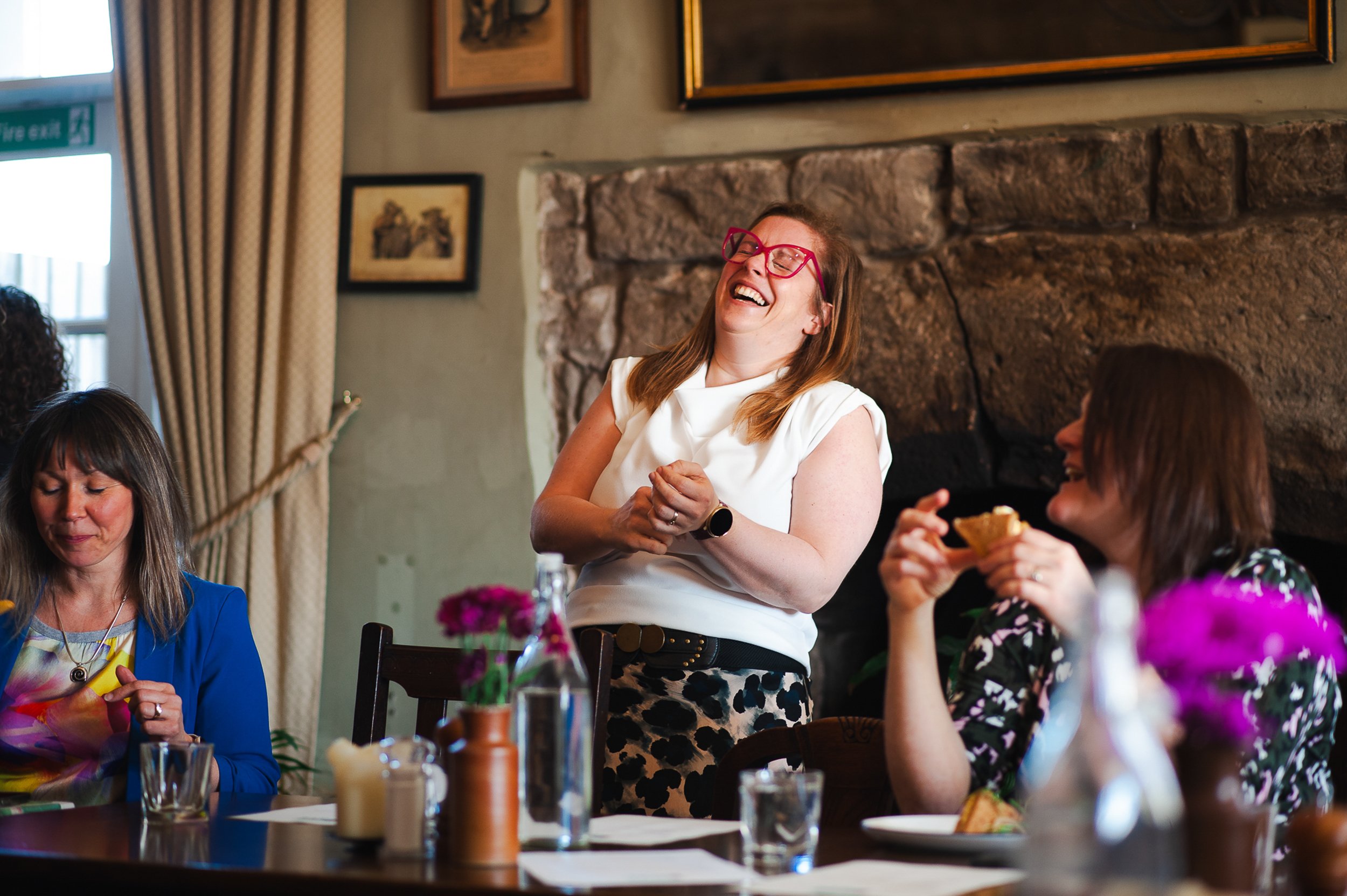 Three women sitting and laughing at a table with drinks and flowers, in a room with stone walls and framed artwork.