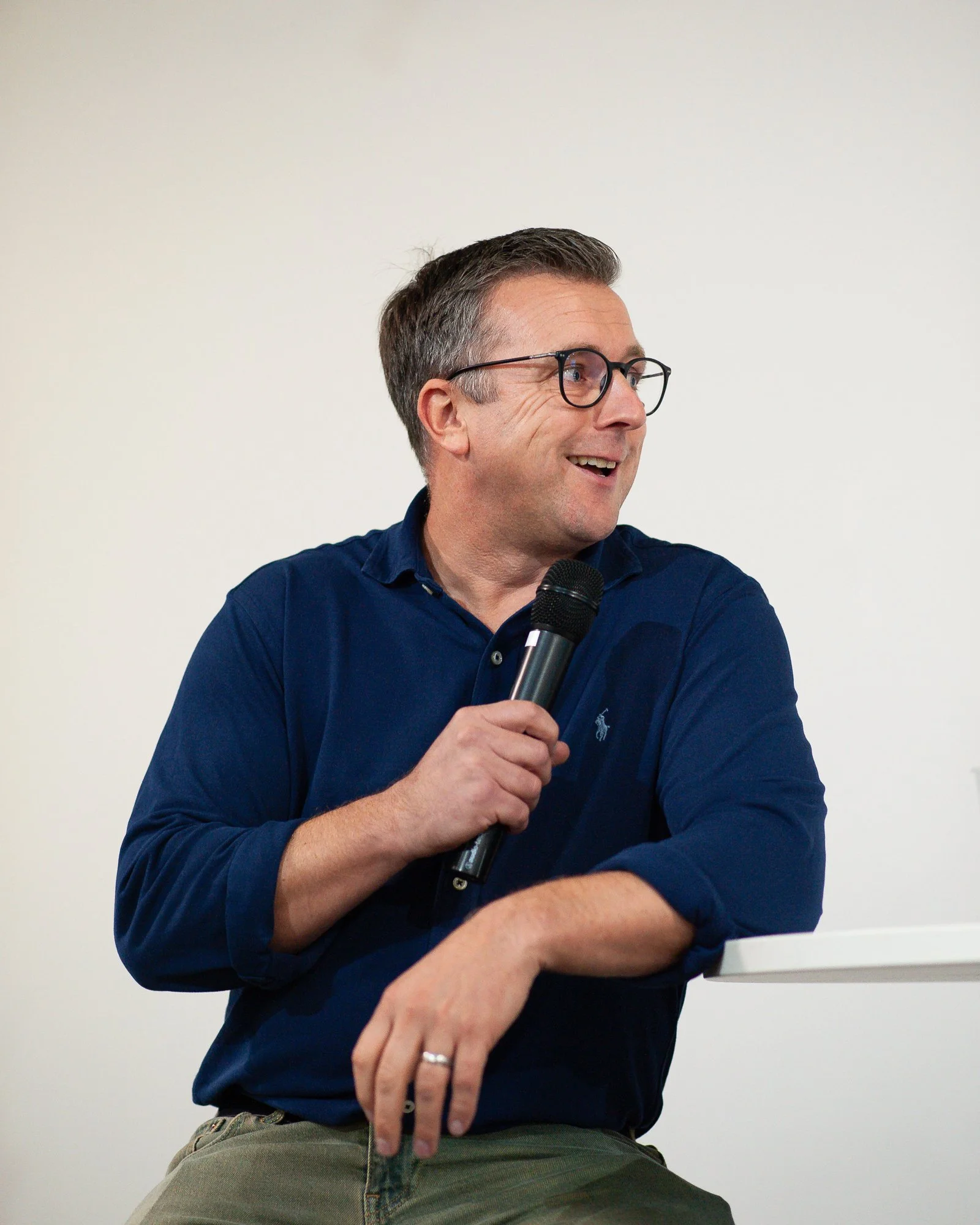 A man sitting and holding a microphone, smiling and looking to his right, wearing glasses, a dark blue shirt with rolled-up sleeves, and olive-green pants.