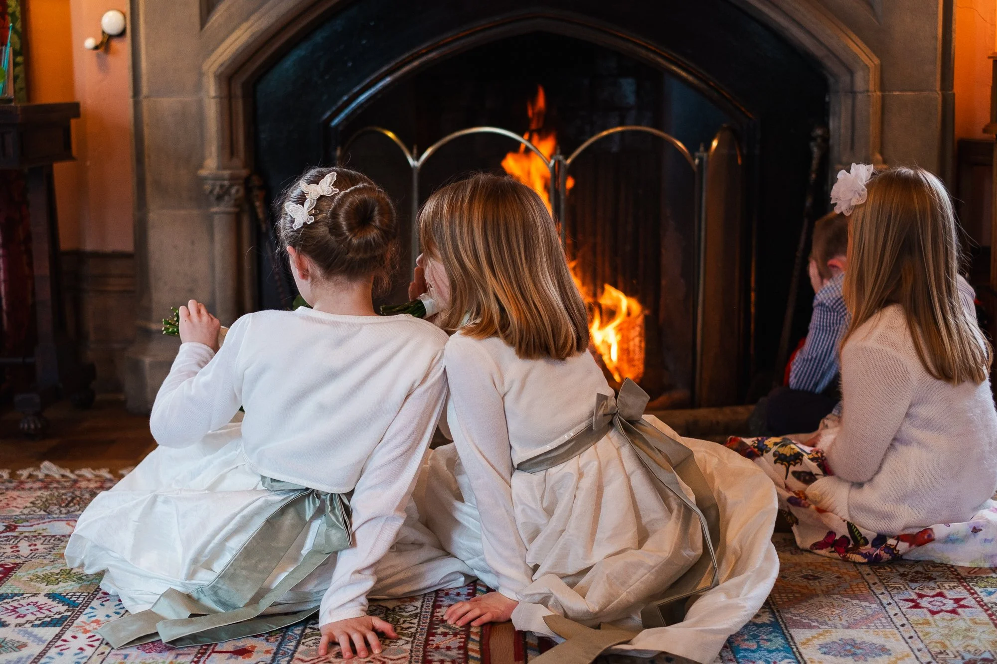Children sitting on a patterned rug in front of a lit fireplace, with two girls whispering to each other.