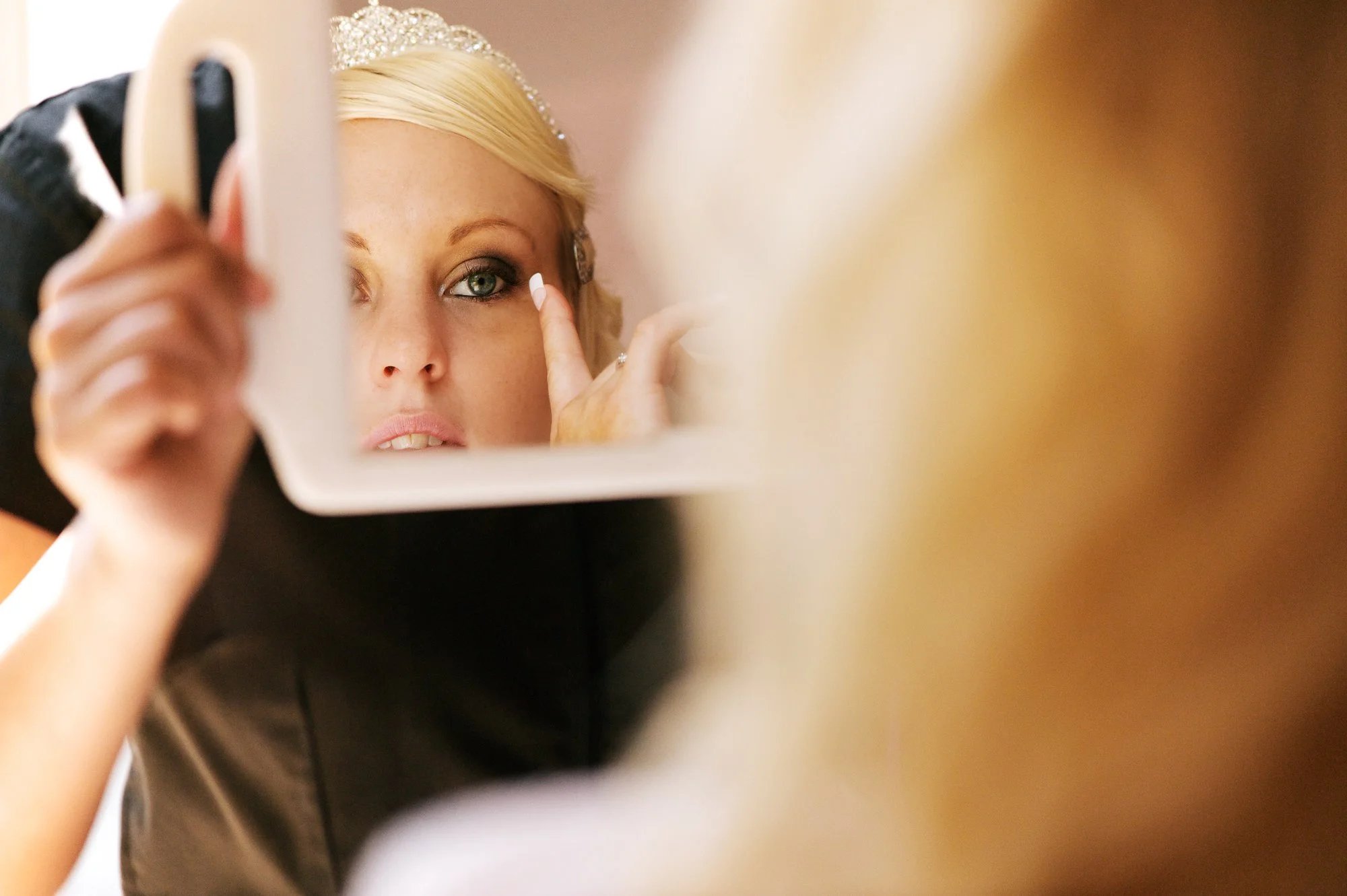 Bride with blonde hair applying makeup while looking in a mirror, wearing a tiara.
