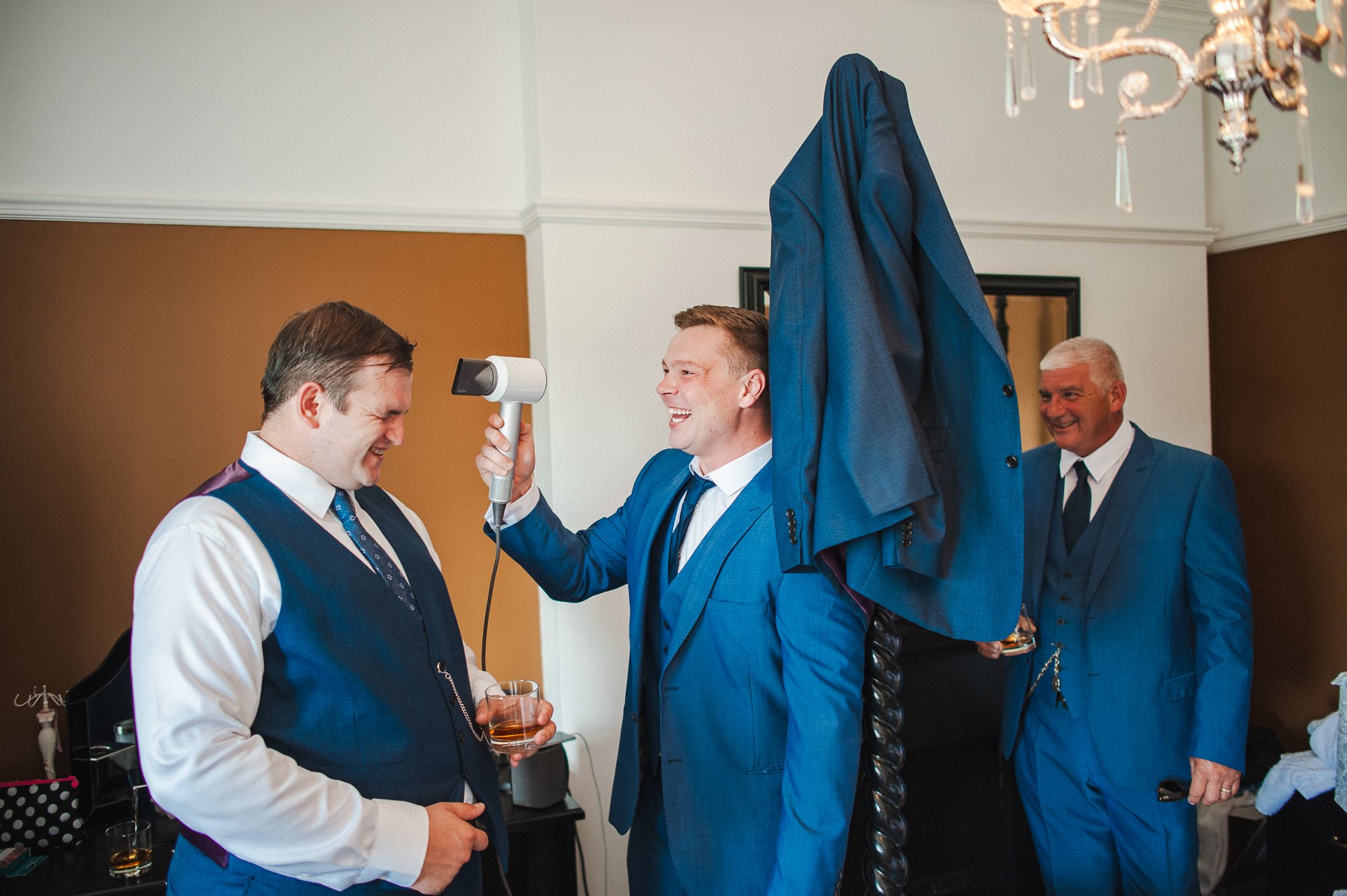 Three men in blue suits laughing and enjoying a moment of fun, with one holding a hairdryer near another's head, in a well-lit room with a chandelier.