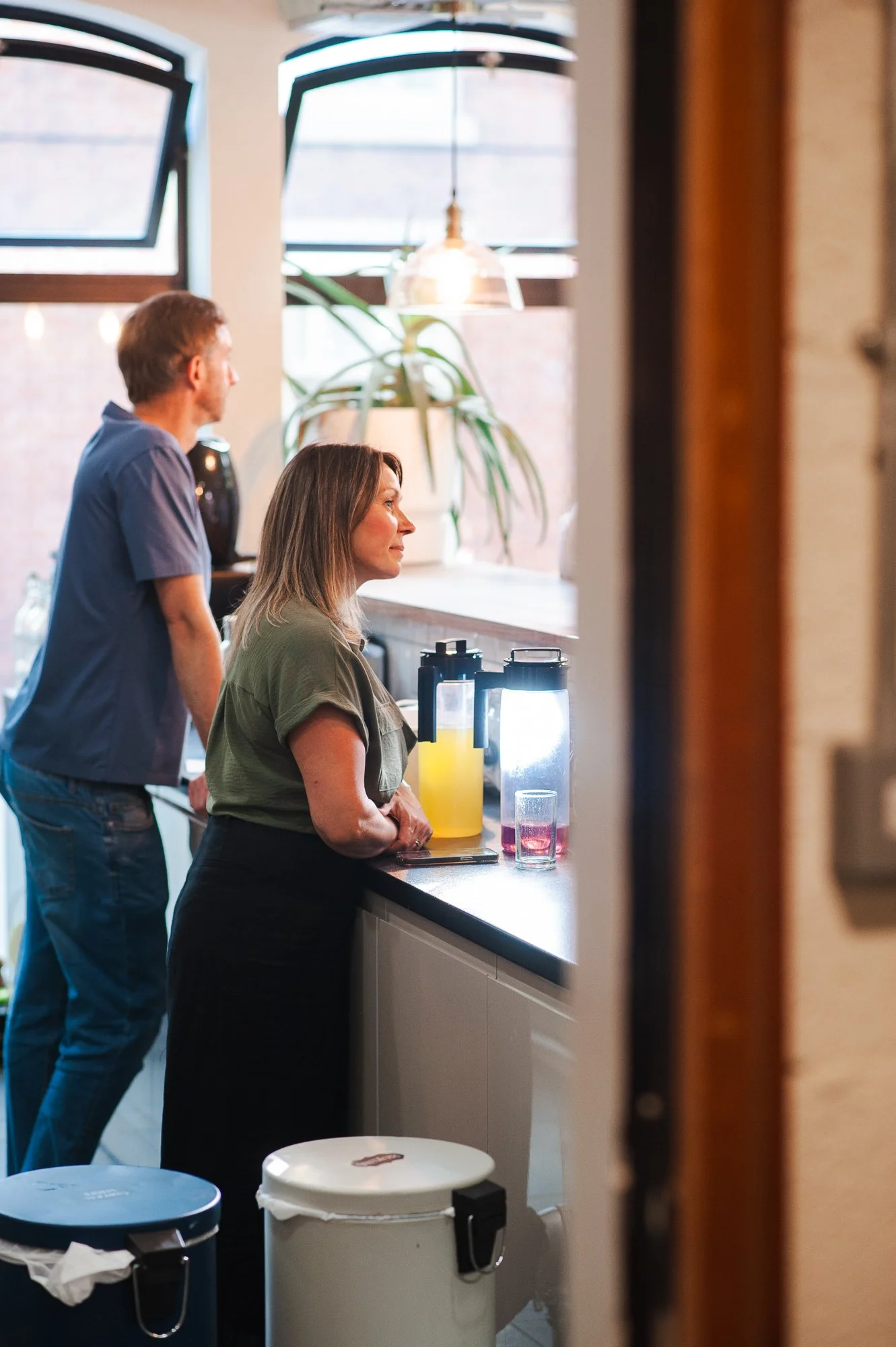 A woman leaning on a kitchen counter with drinks and pitchers, while a man stands behind her gazing out the window, in a well-lit modern kitchen.
