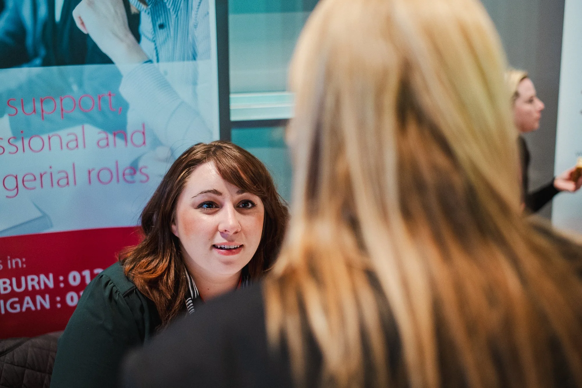 A woman with brown hair sitting at a table, engaging in a conversation with a woman with blonde hair. In the background, there is a banner with pink and white text.