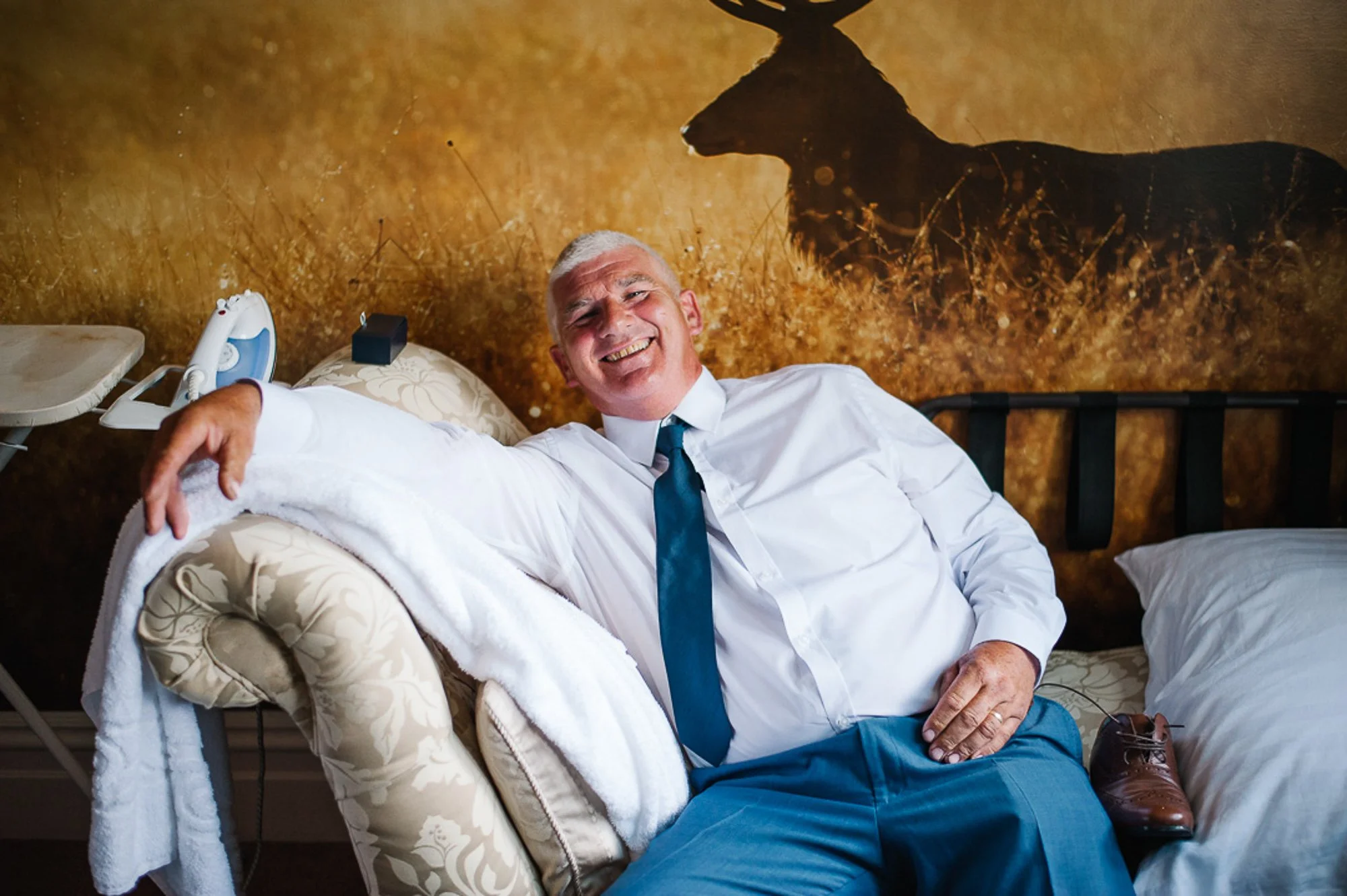 An older man with gray hair and a big smile reclining on a beige floral armchair in a room with a large mural of a moose on the wall behind him. He is wearing a white shirt, blue tie, and blue pants, with brown shoes on the bed beside him. There is a