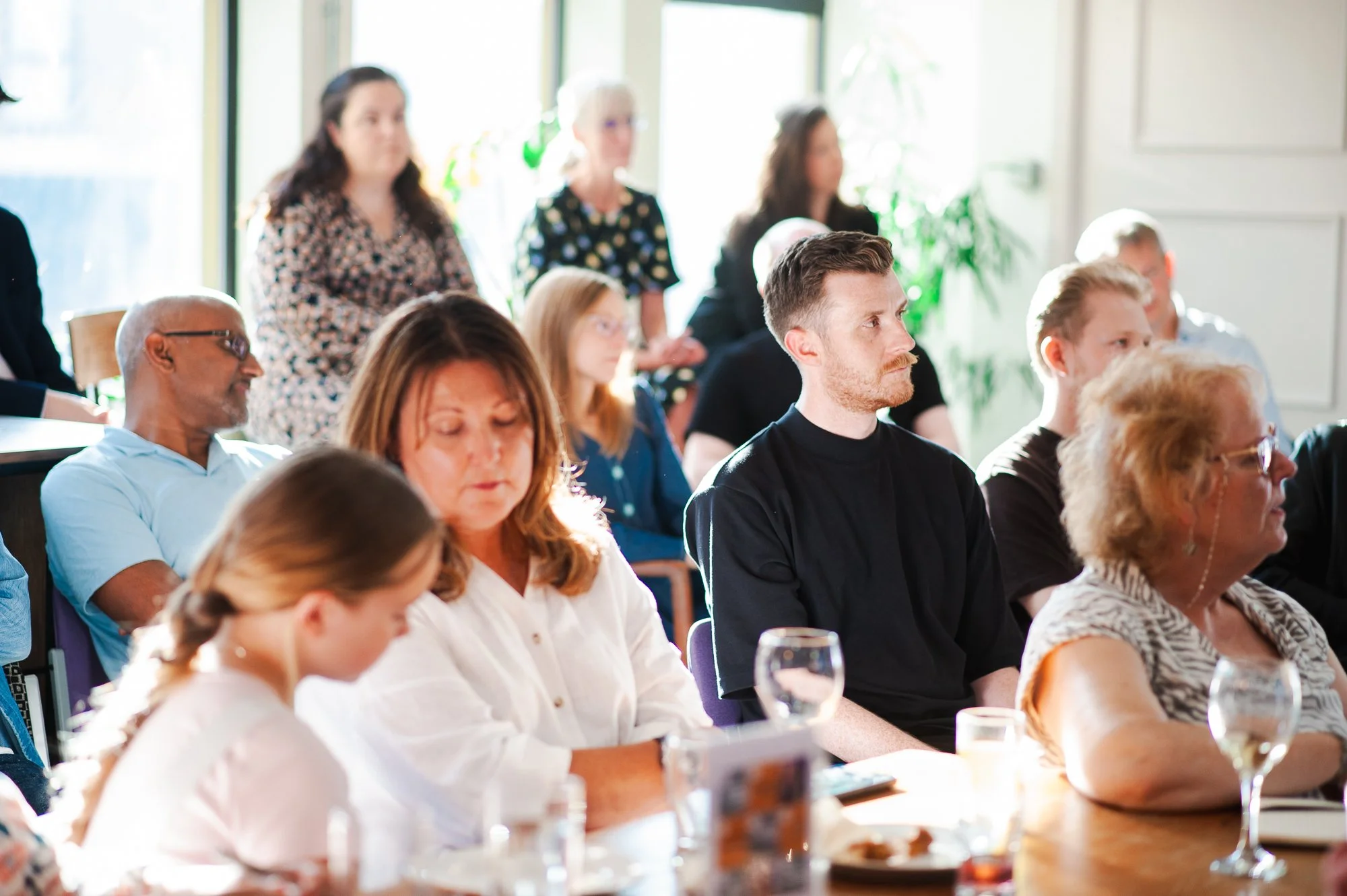 People attending a meeting or seminar, sitting and listening attentively in a bright room with large windows and green plants.