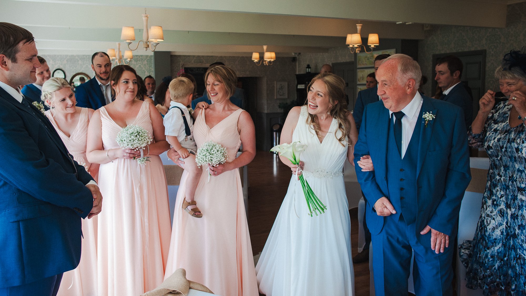 Wedding celebration with bride holding lilies, surrounded by family and friends in a warmly lit room.