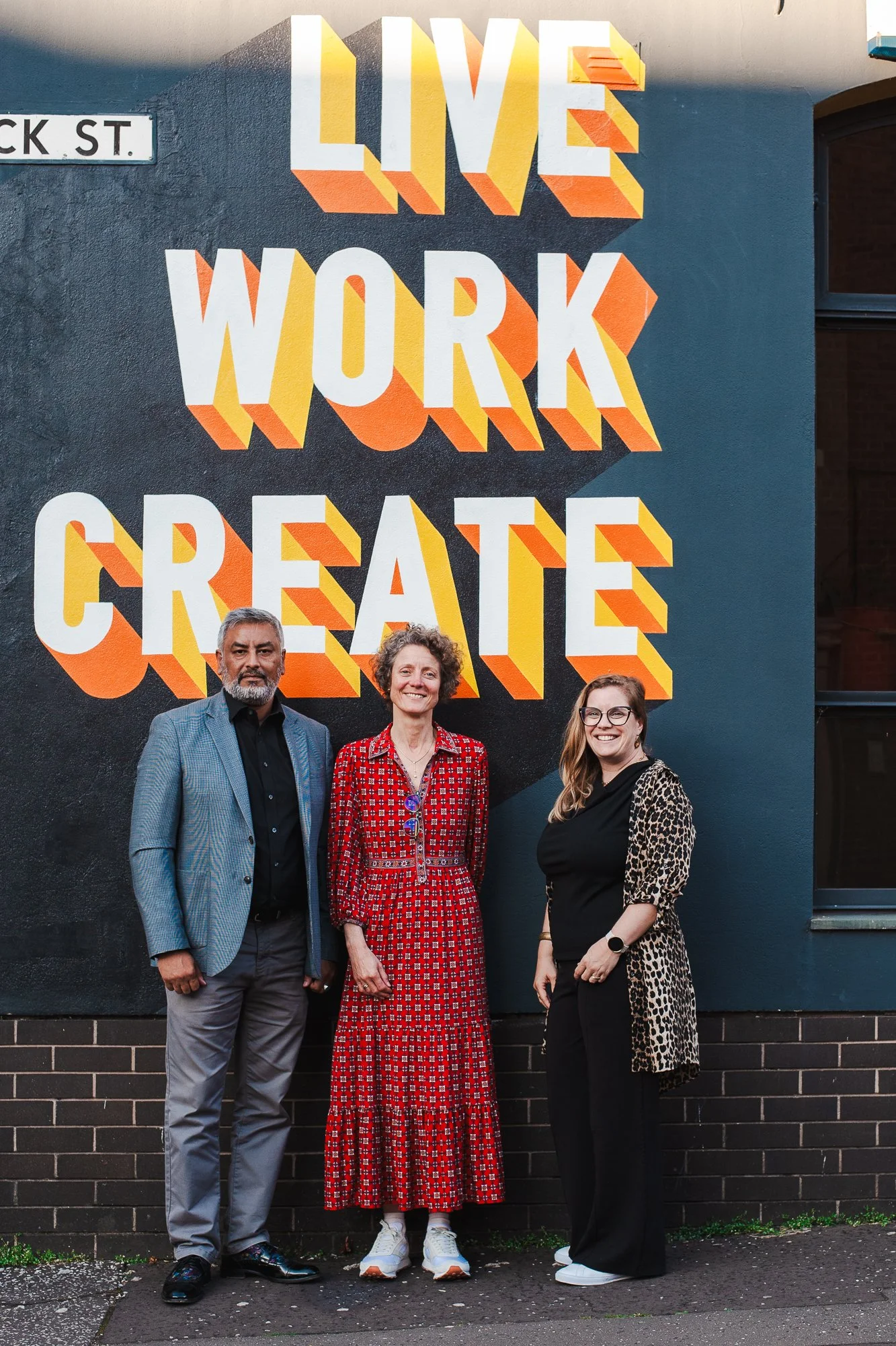 Three people standing in front of a mural that reads 'LIVE WORK CREATE' in large, colorful 3D letters. A man in a gray checkered blazer, a woman in a red patterned dress, and another woman in black with leopard print sleeves are smiling.