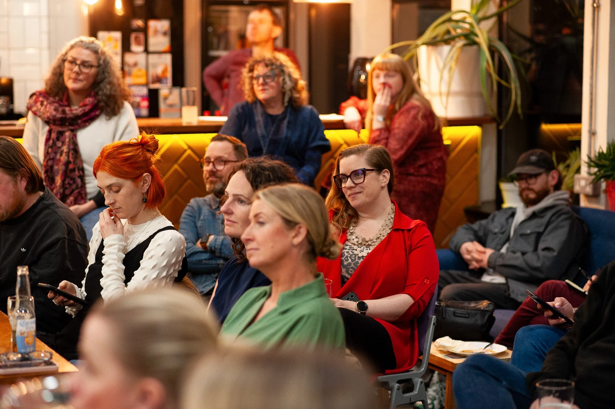 Group of people attending a presentation or event in a cozy indoor setting, some seated and some standing, with warm lighting and decor including plants and framed pictures.