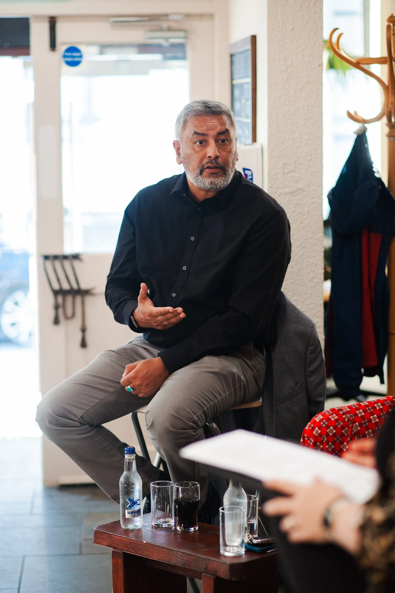 A man with gray hair and a beard, dressed in a black shirt, is sitting on a high chair in a casual indoor setting, talking or explaining something to a group. In front of him on a small table are a bottle of water, a glass of dark soda, and some empt