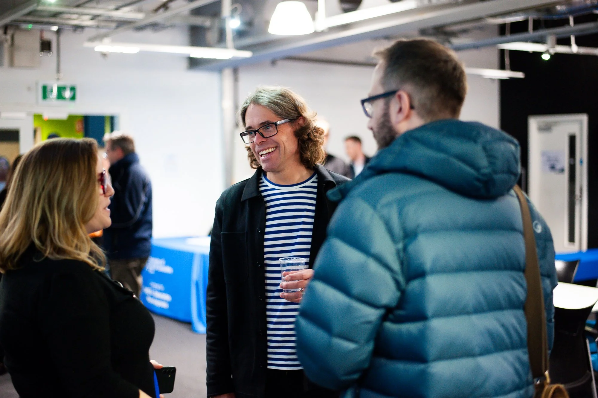 Three people engaged in conversation at an indoor event, with a background of a blue table and other attendees.