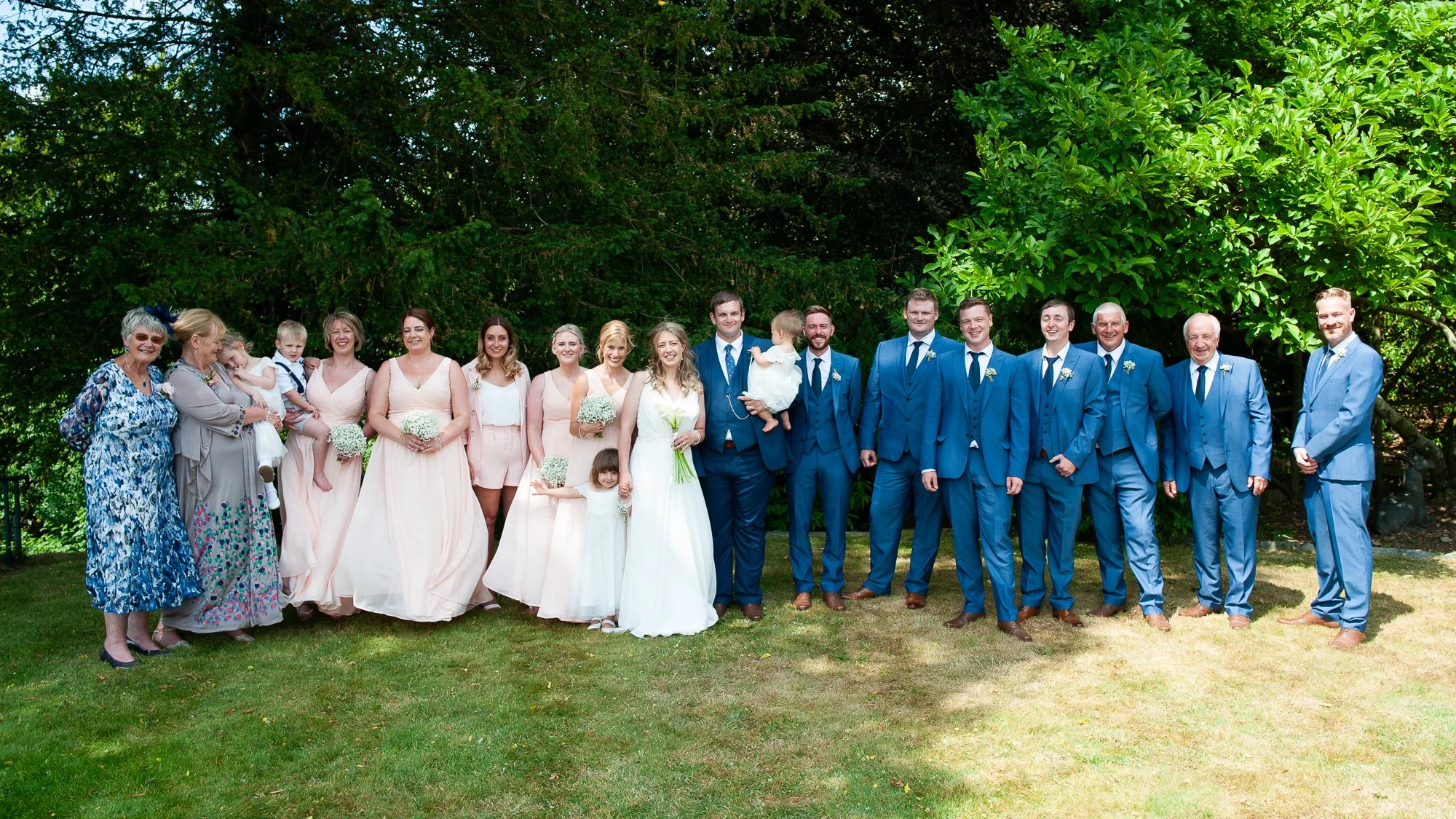 Group of wedding guests, including bridesmaids, groomsmen, and family, standing outdoors on grass with lush green trees in the background.
