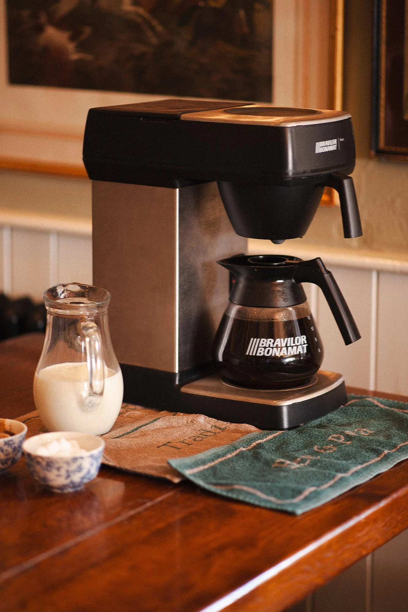 A coffee brewing station with a drip coffee maker, a glass carafe filled with brewed coffee, a small pitcher of milk, and small bowls on a wooden table.