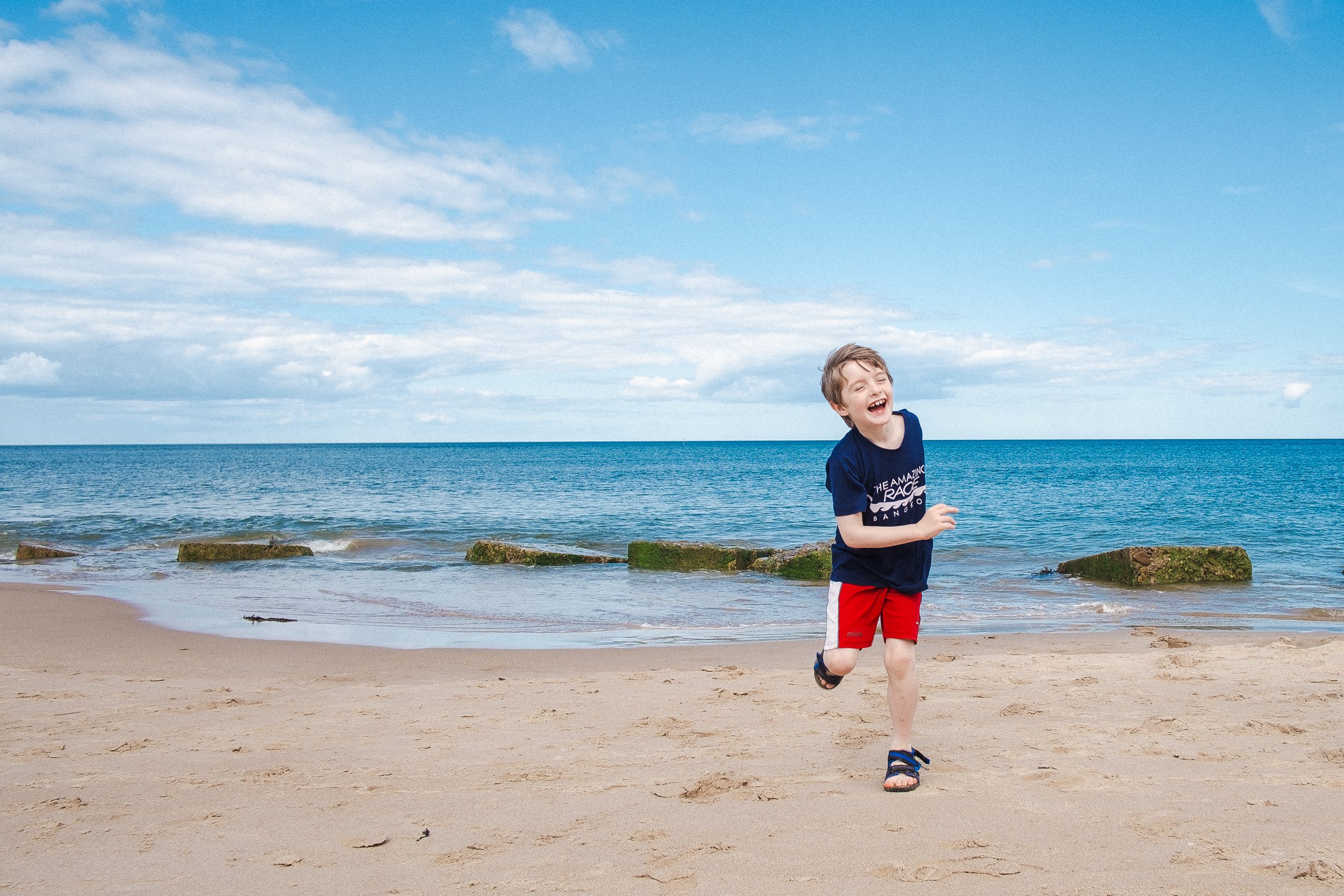 Child running and laughing on a sandy beach with blue ocean and sky in the background.