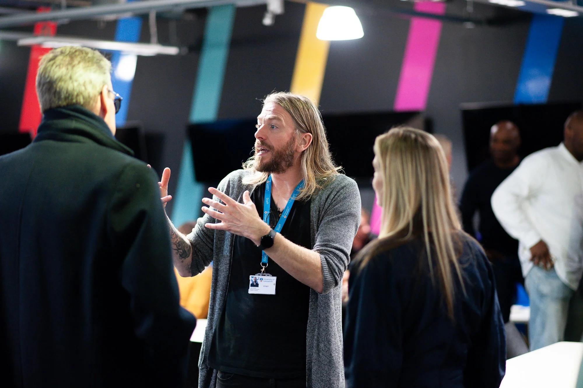 A man with long hair and beard gesturing and talking to two people at a conference or event, with a group of people in the background.