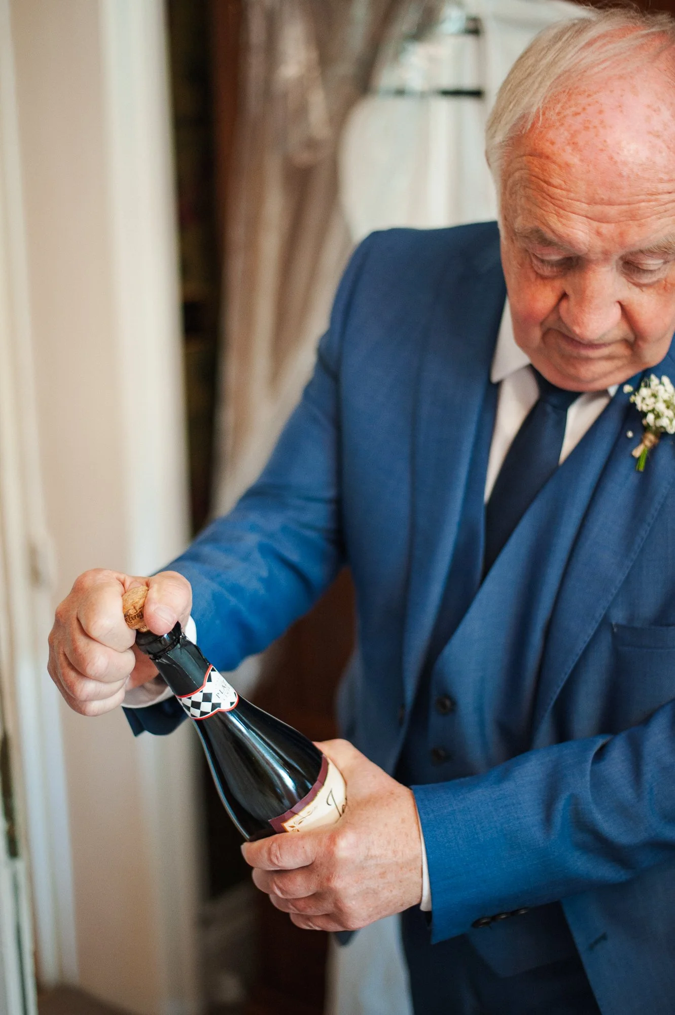 An elderly man in a blue suit opening a bottle of champagne.
