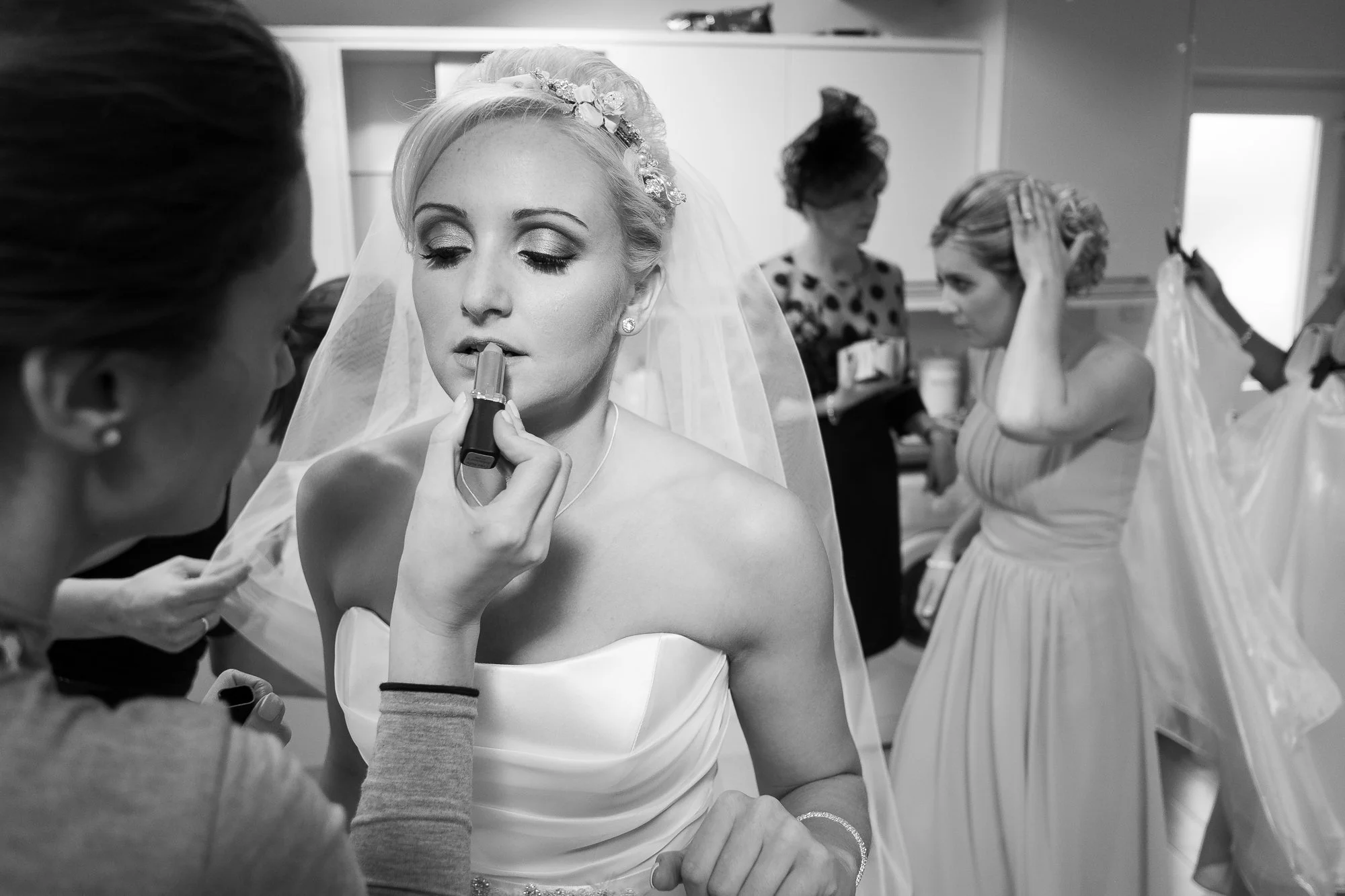 A bride getting her makeup done on her wedding day while a makeup artist applies lipstick, with other women preparing in the background.