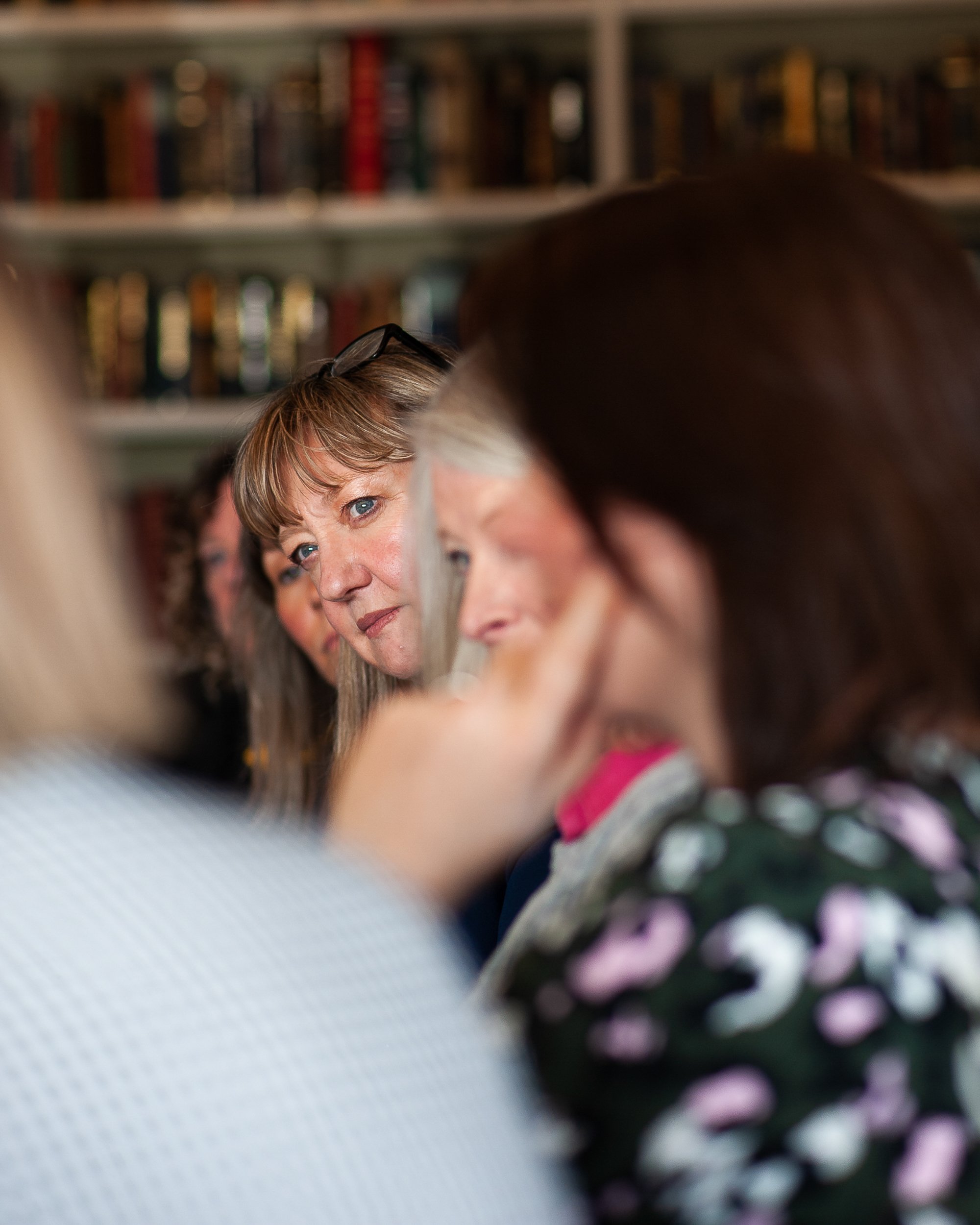 A group of people in a library setting, with one woman in focus looking intently. Bookshelves are visible in the background.