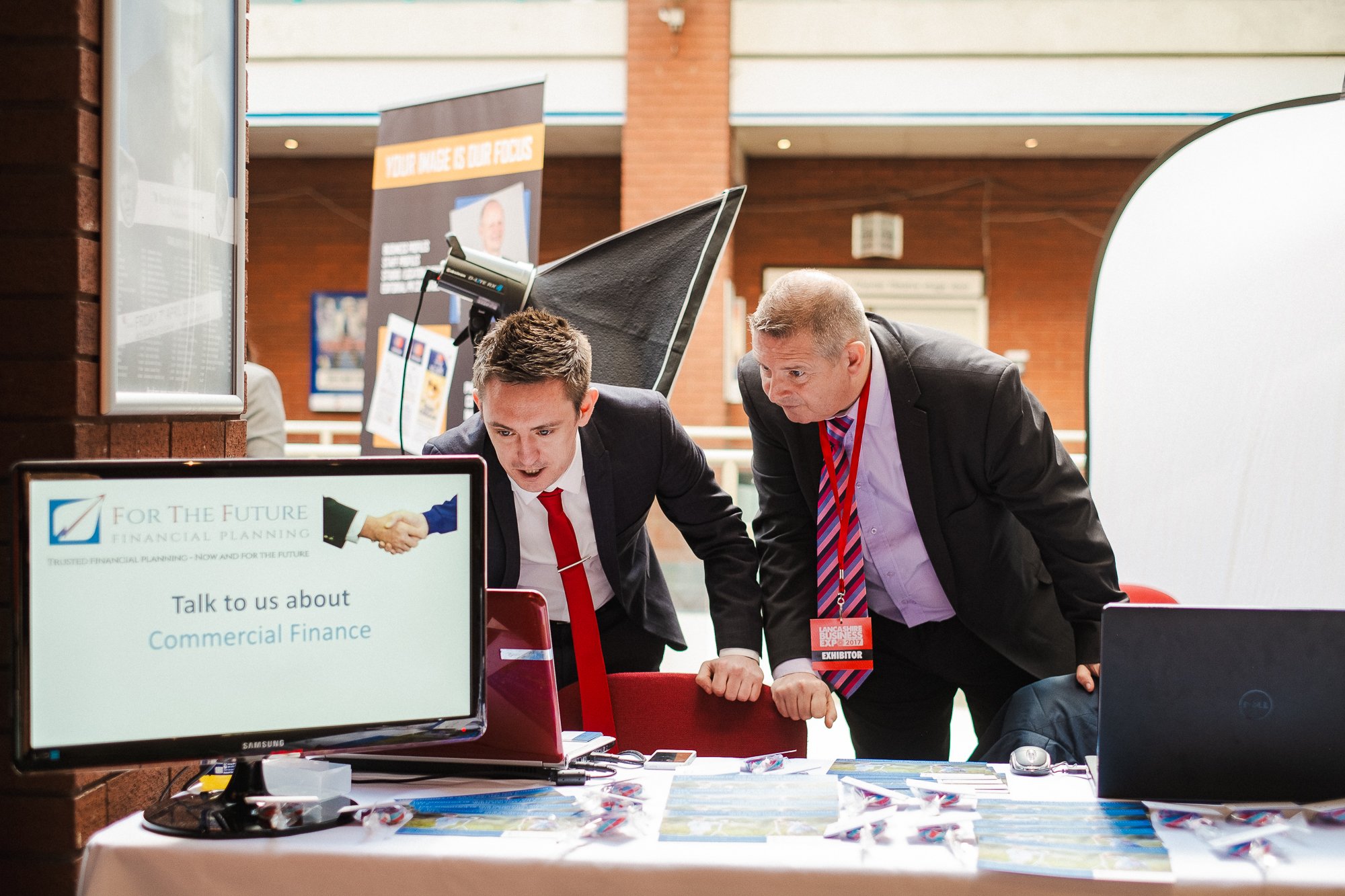 Two men in business suits are looking at a computer screen at a booth about commercial finance, with promotional materials and a Dell laptop on the table.