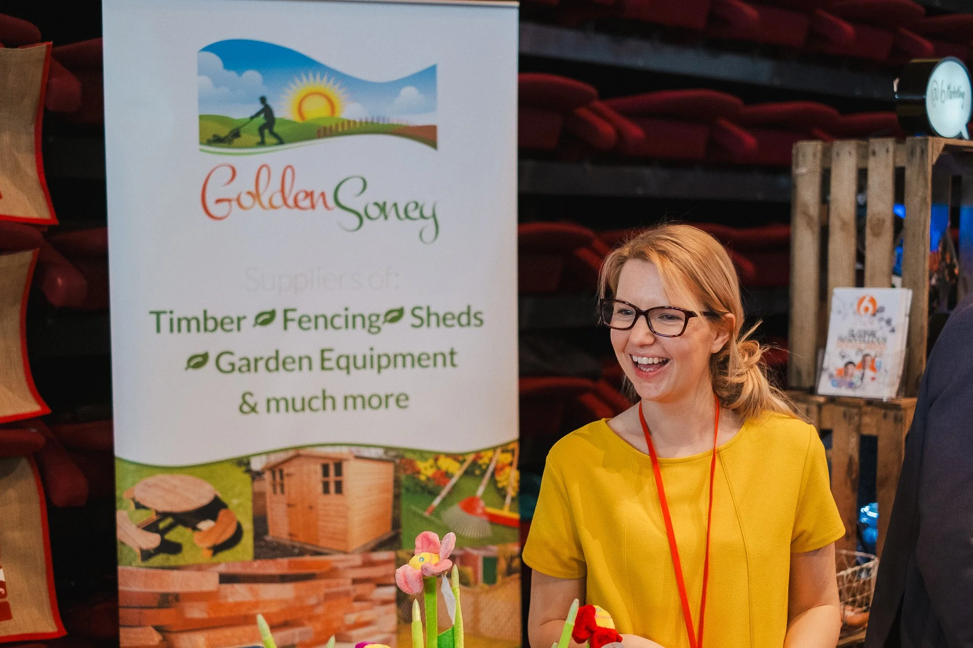 A woman with glasses and a yellow shirt smiling at a booth with a sign for Golden Sawyer, a supplier of timber, fencing, sheds, garden equipment, and more. There are colorful garden decorations and wooden sheds around her.