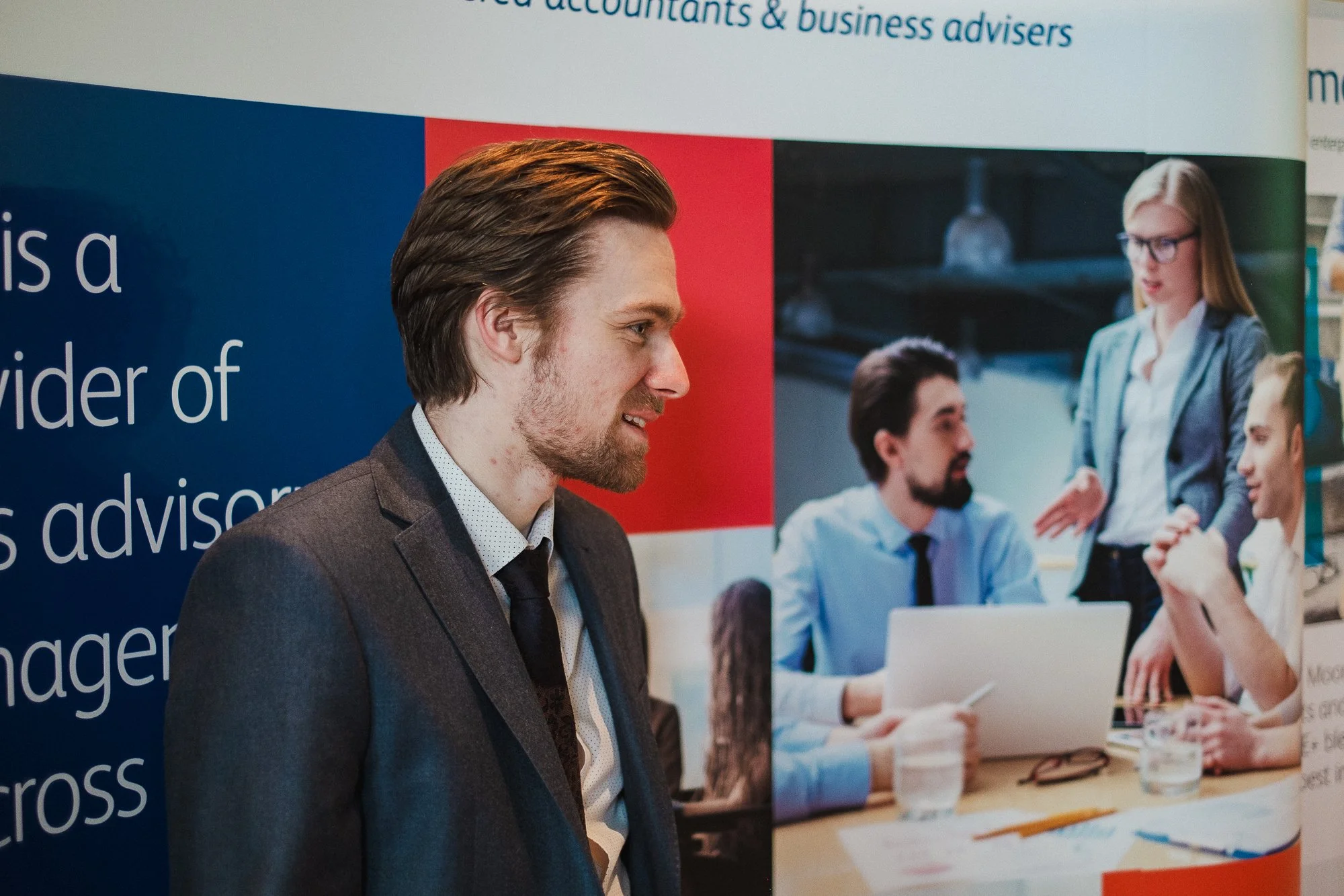 A man in a dark suit standing in front of a poster or display with pictures of people in a business meeting, with a woman talking to two men sitting at a table with laptops and glasses of water.