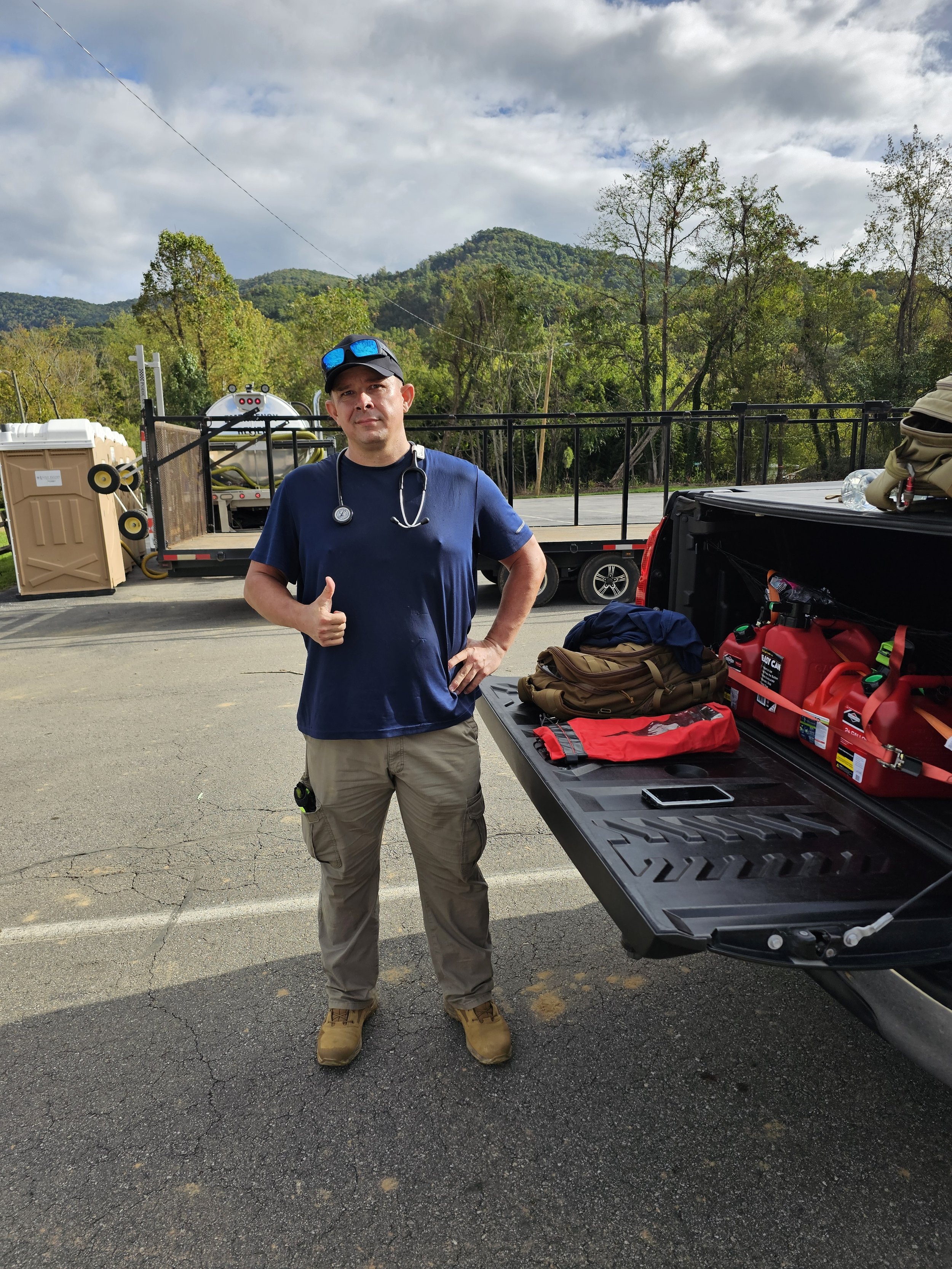 A man wearing a navy blue t-shirt, khaki cargo pants, tan boots, a black cap with blue sunglasses on top, and a stethoscope around his neck standing next to a utility vehicle with emergency equipment and supplies, giving a thumbs-up with one hand. Th