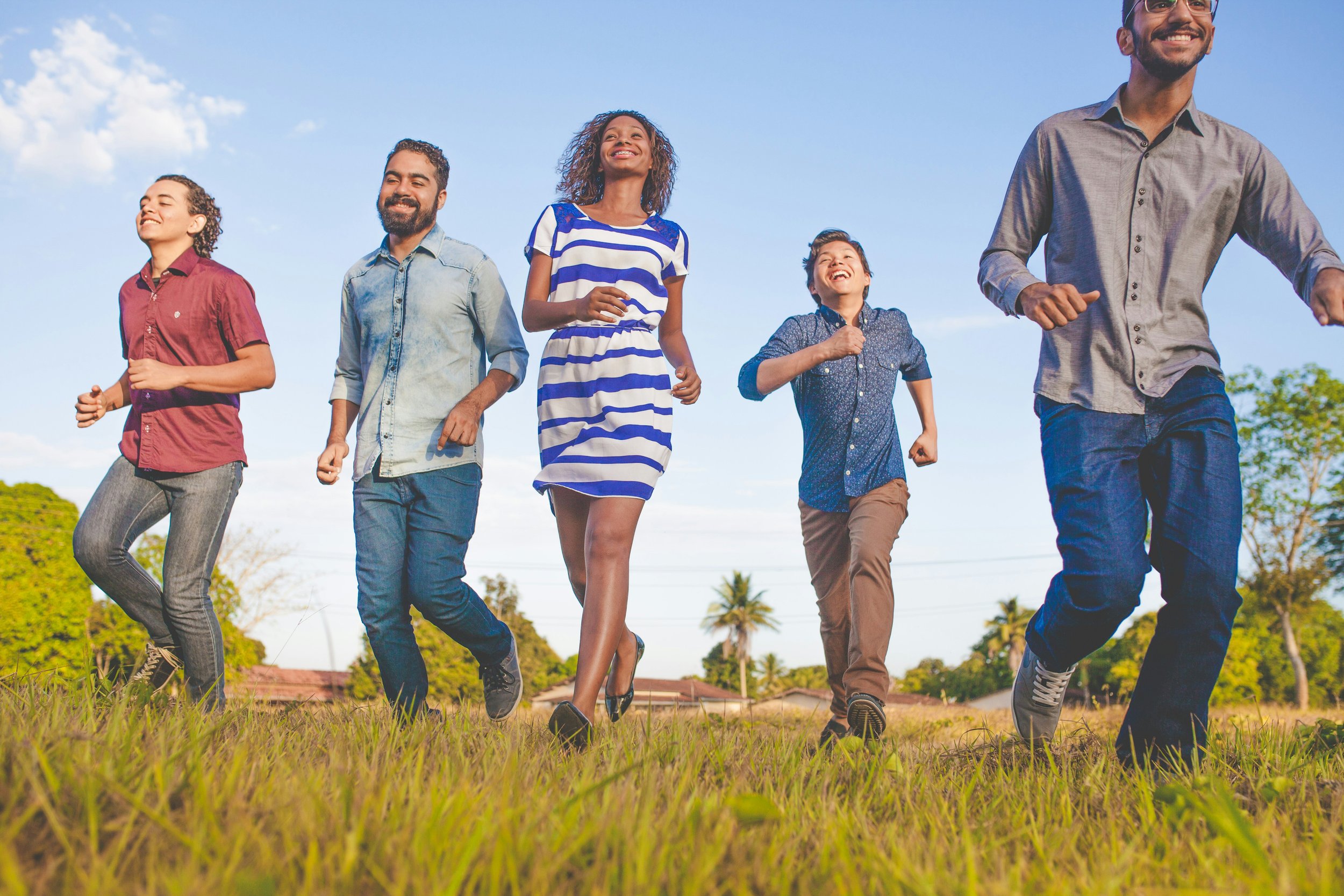 A group of five diverse young adults smiling and running in a grassy field on a sunny day