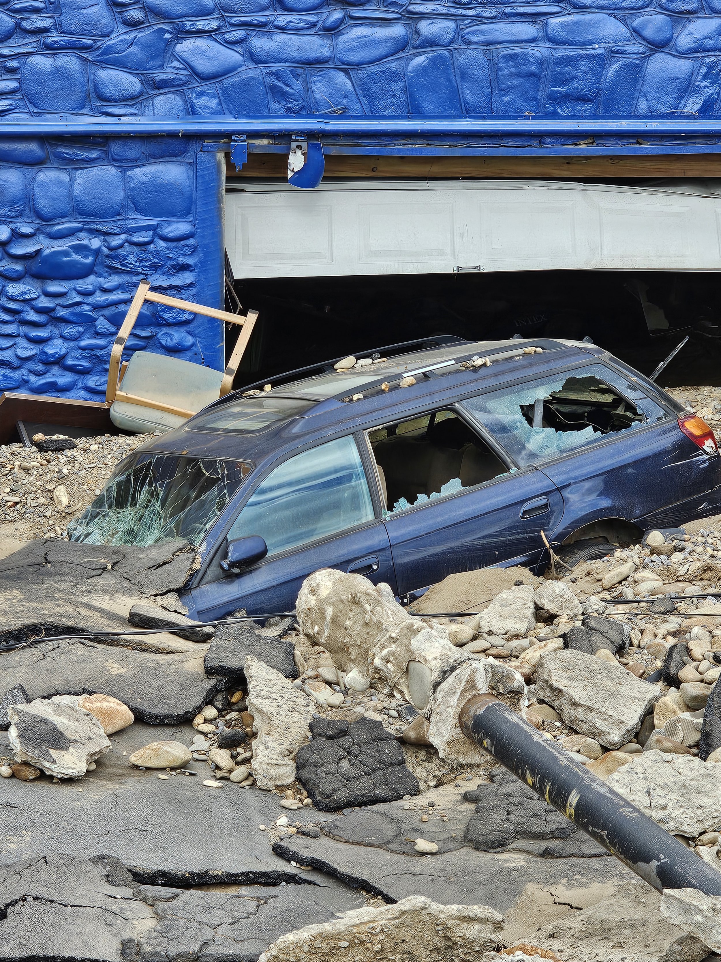 A car has crashed into a building, breaking through a garage door and being partially buried under debris and large rocks. The car's windows are shattered, and the scene shows significant destruction.