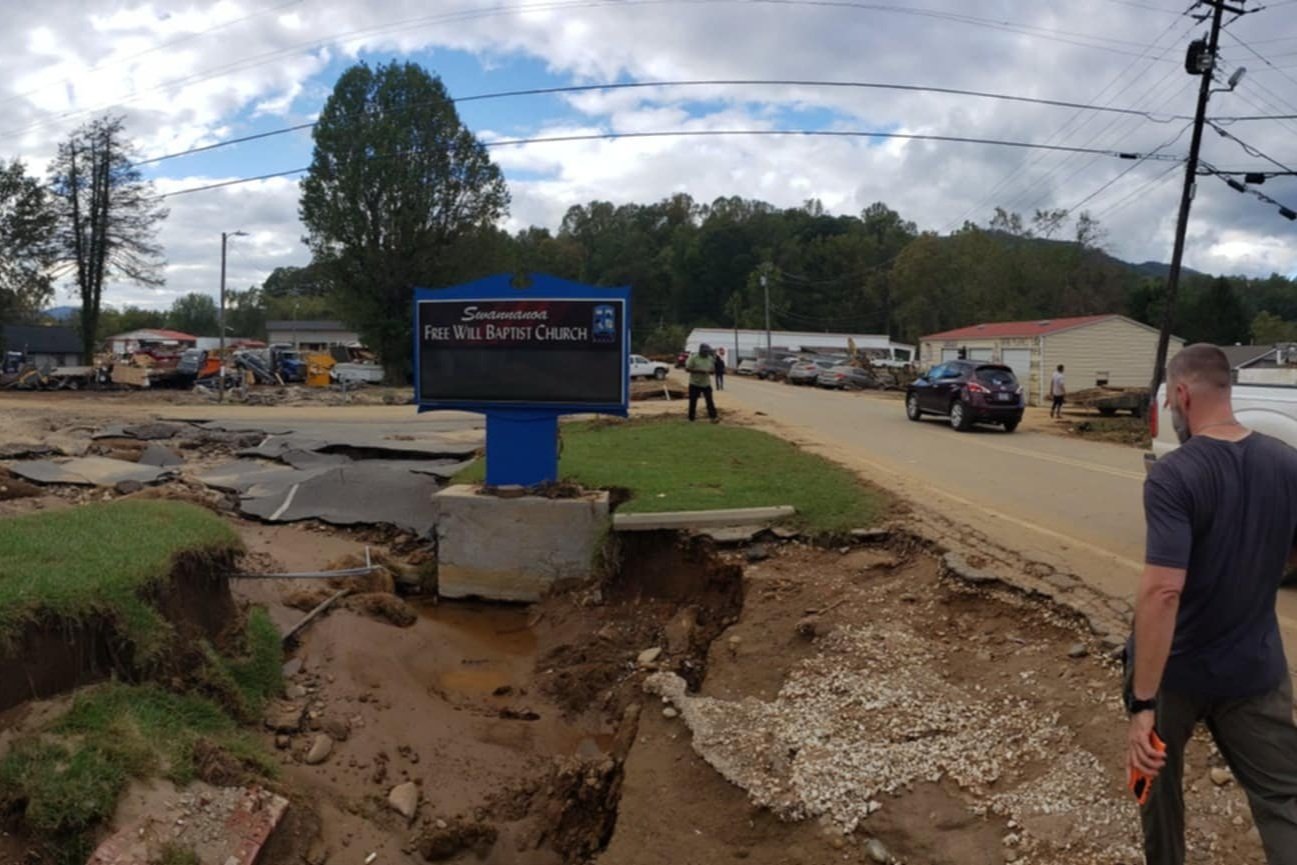 Damaged sidewalk and road with exposed dirt and gravel, construction or repair work in progress, vehicles parked and driving on the street, trees and buildings in the background, and a man looking at the scene.