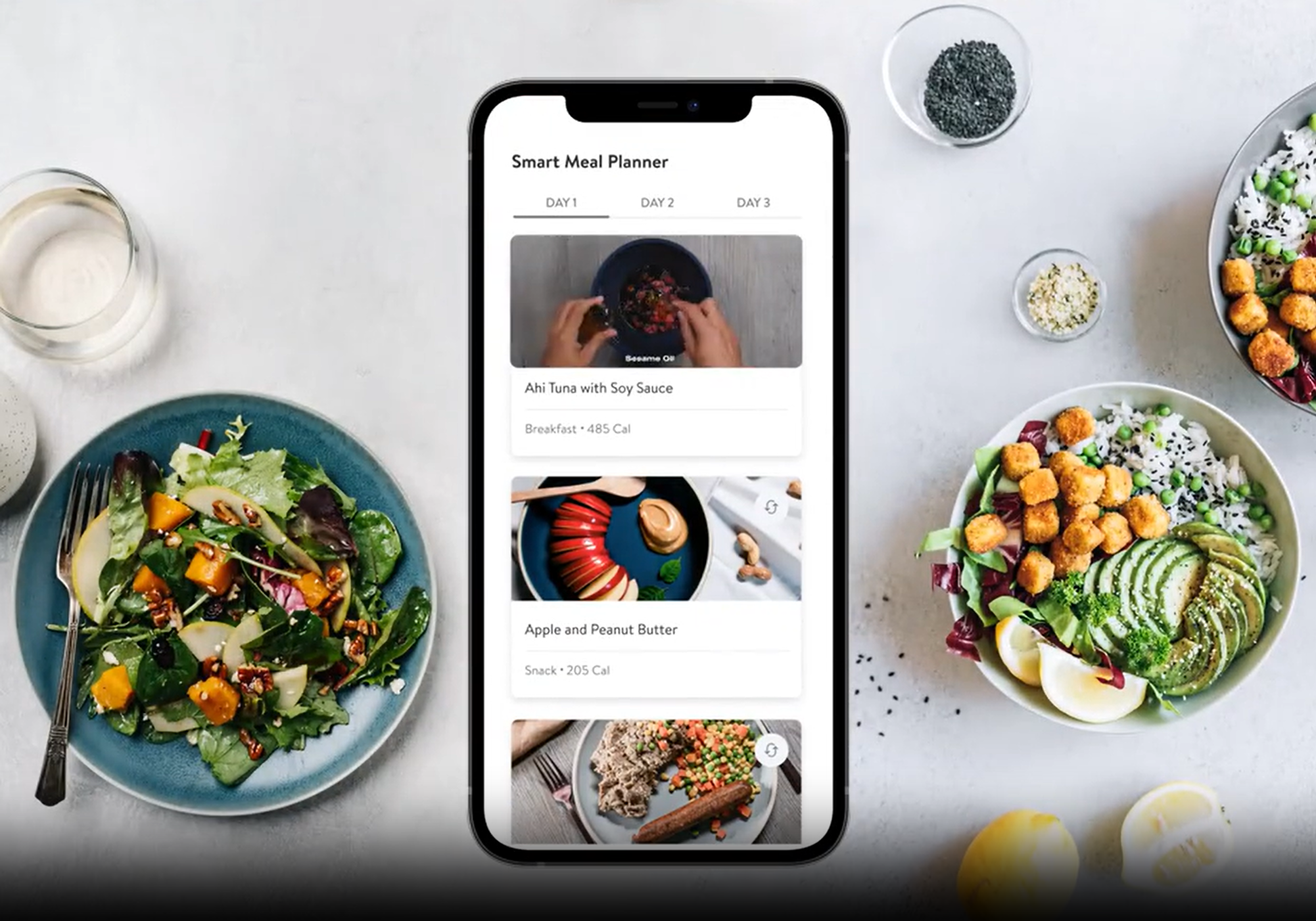 A smartphone displaying a meal planner app with three meal options on a white surface. Surrounding the phone are plates of colorful salads, bowls of salad toppings, and glasses of drinks.