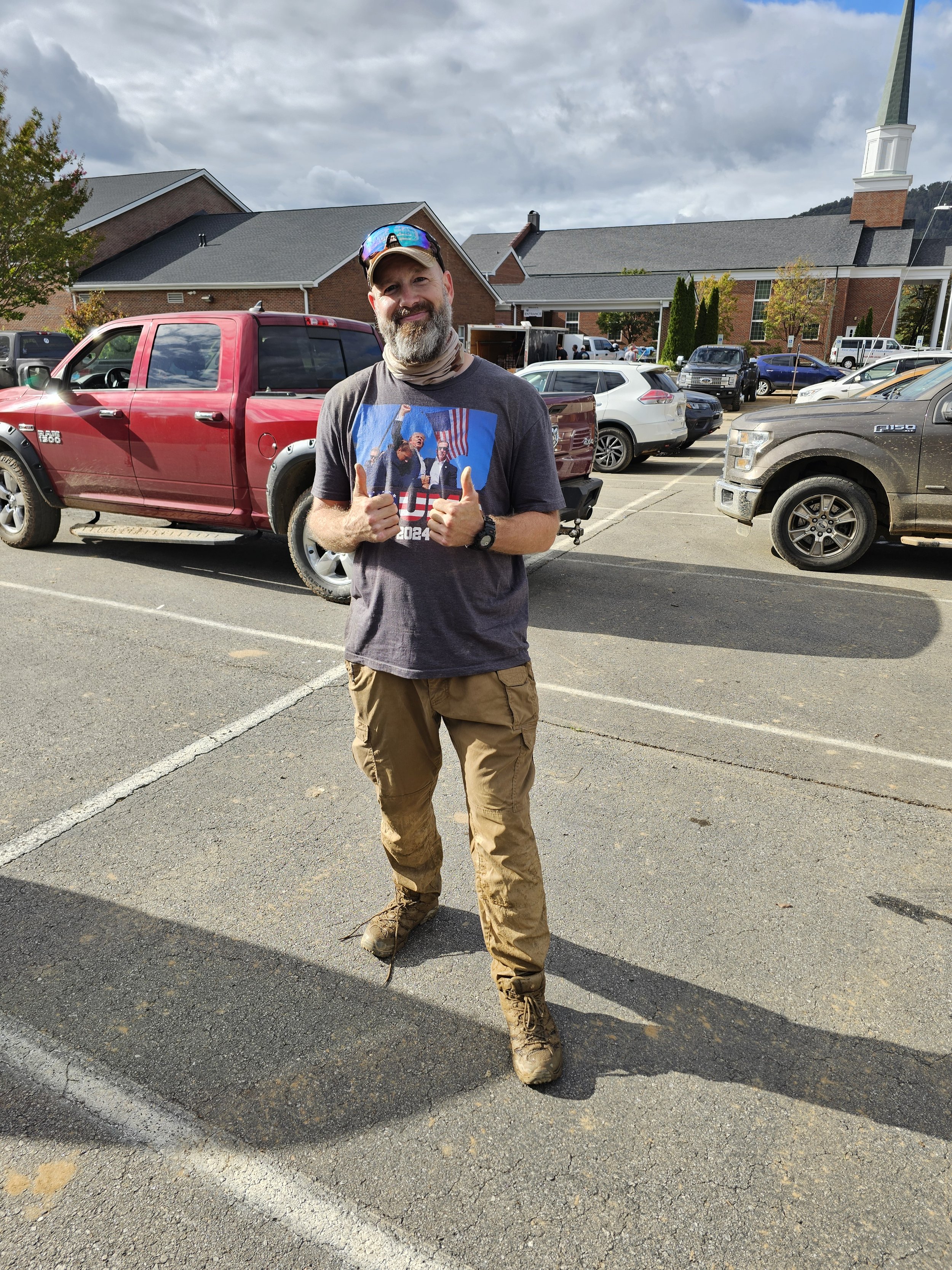 A man with a beard and sunglasses giving a thumbs up in a parking lot, wearing a dark graphic t-shirt, tan cargo pants, and hiking boots, with cars and a church in the background.
