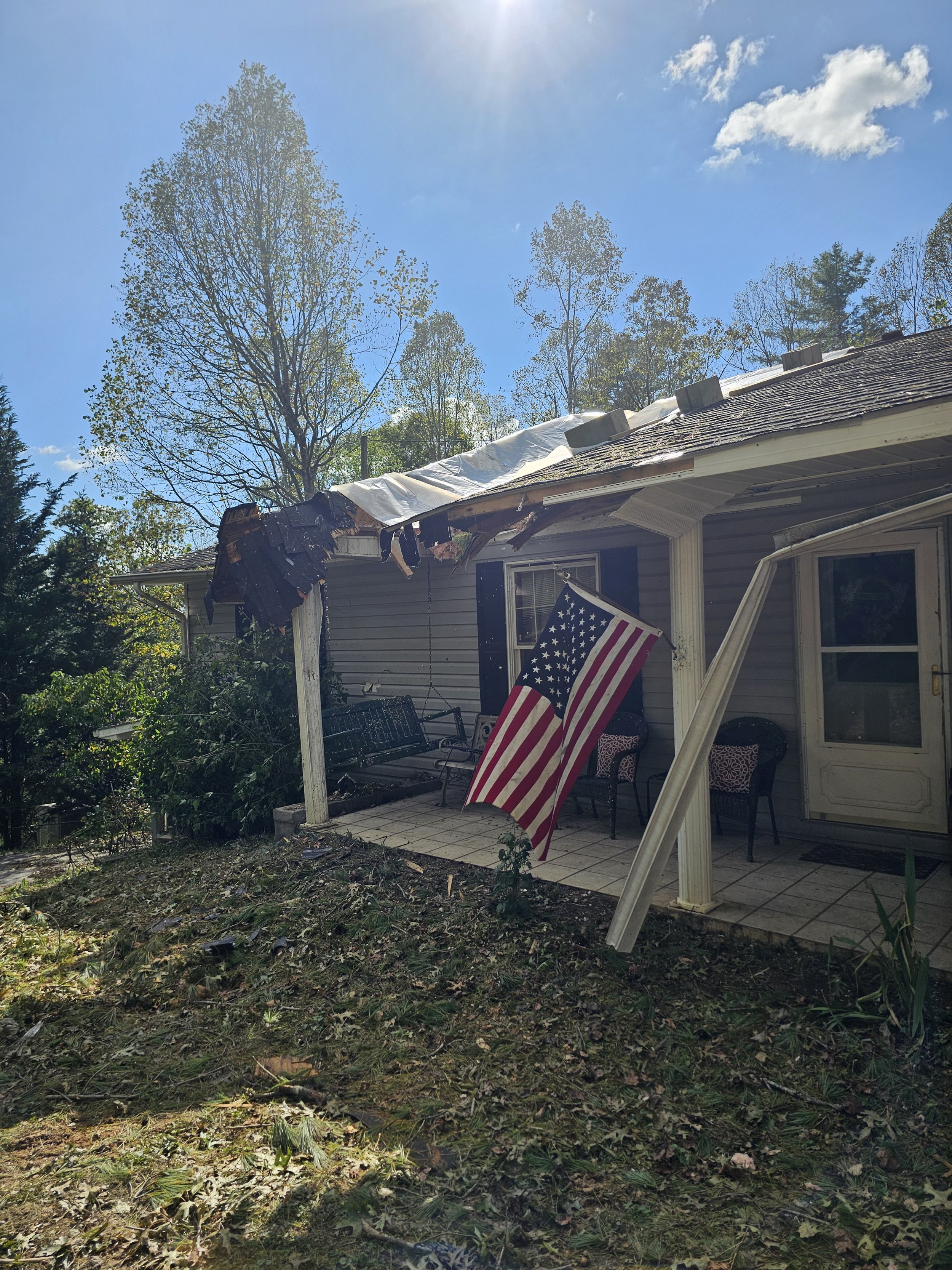 Damaged house with a roof in disrepair, American flag hanging outside, patio with chairs, and debris on the yard.