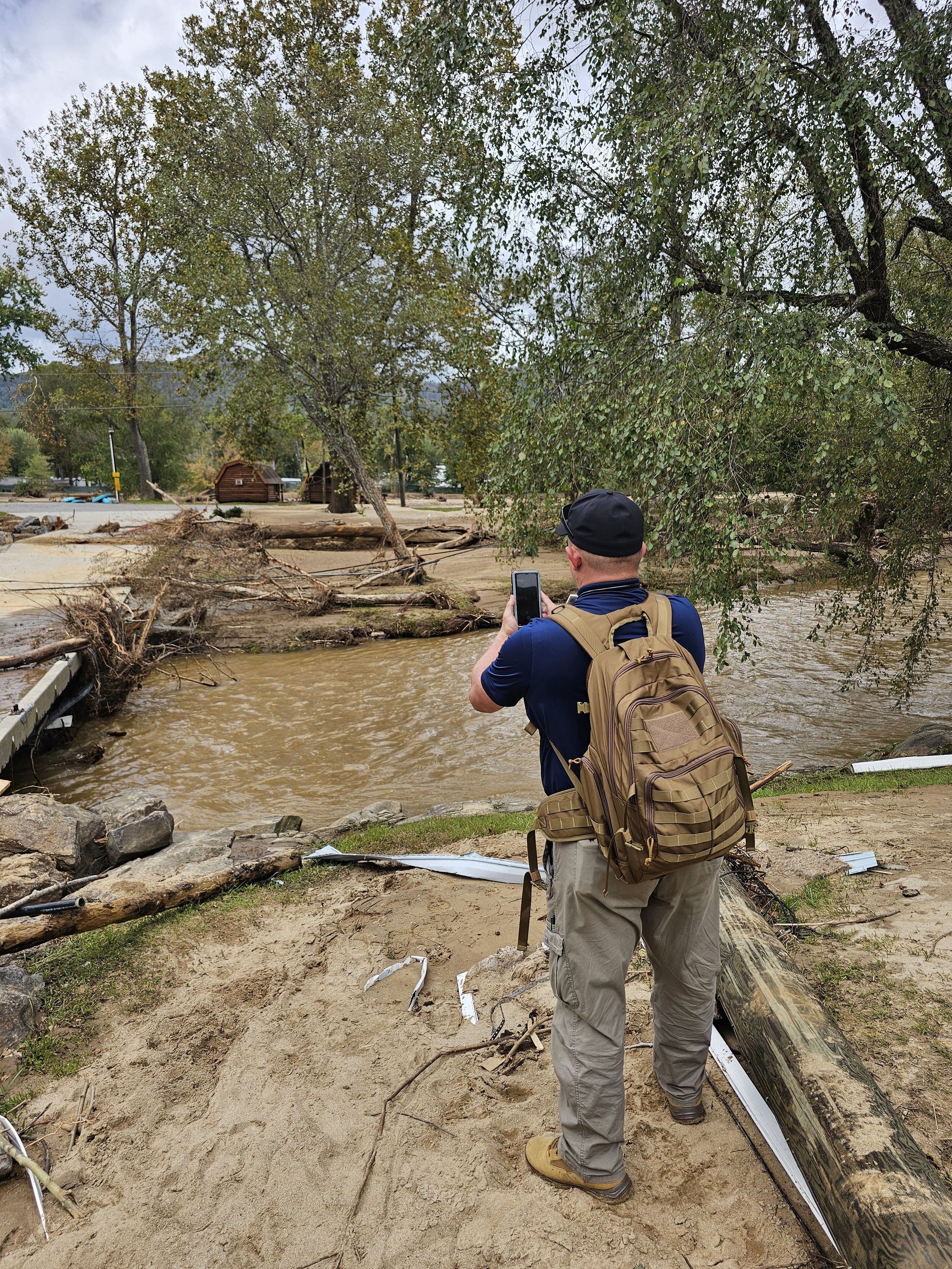 A man with a backpack taking a photo of flood damage, with fallen trees and debris in a muddy water landscape, under an overcast sky.