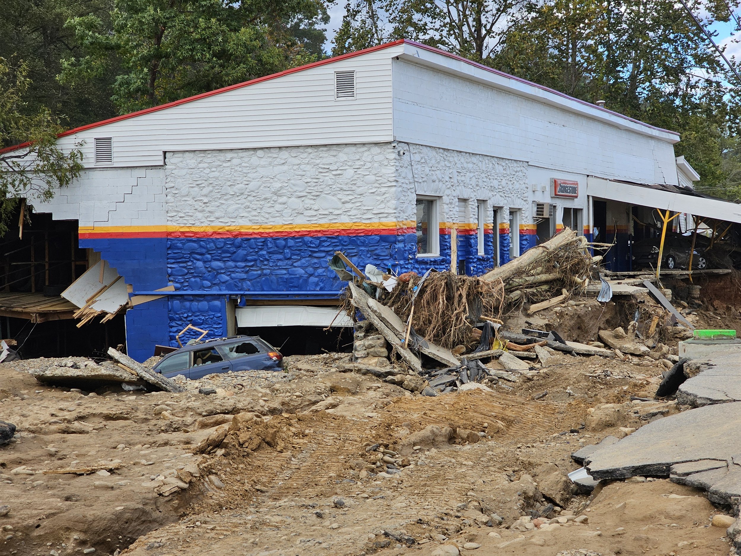 A building with part of its structure damaged and debris scattered around, including a fallen tree and a damaged car. The building shows signs of severe destruction, likely caused by a natural disaster.