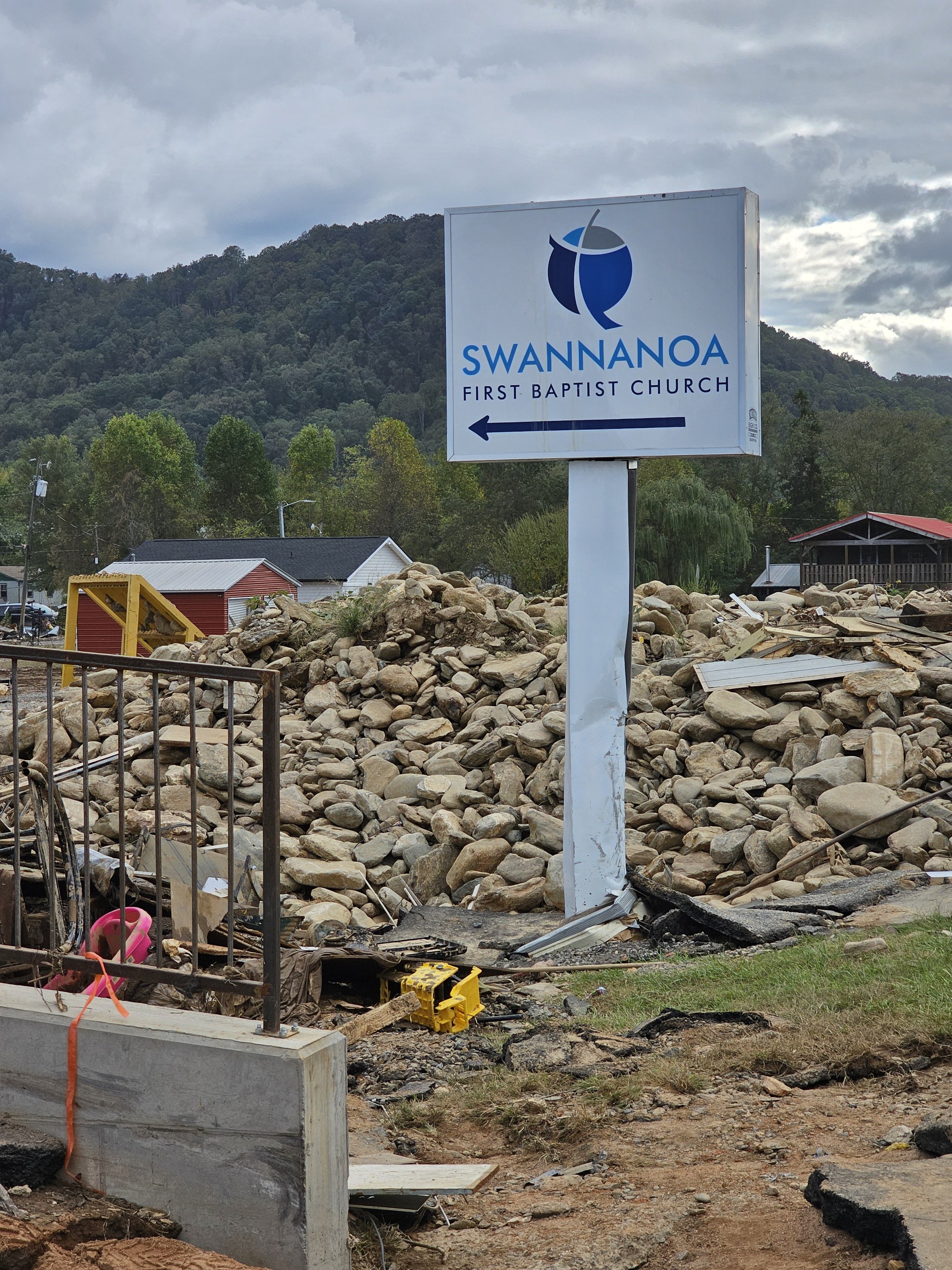 A damaged sign for Swannanoa First Baptist Church with debris and rocks around, and a mountain in the background.