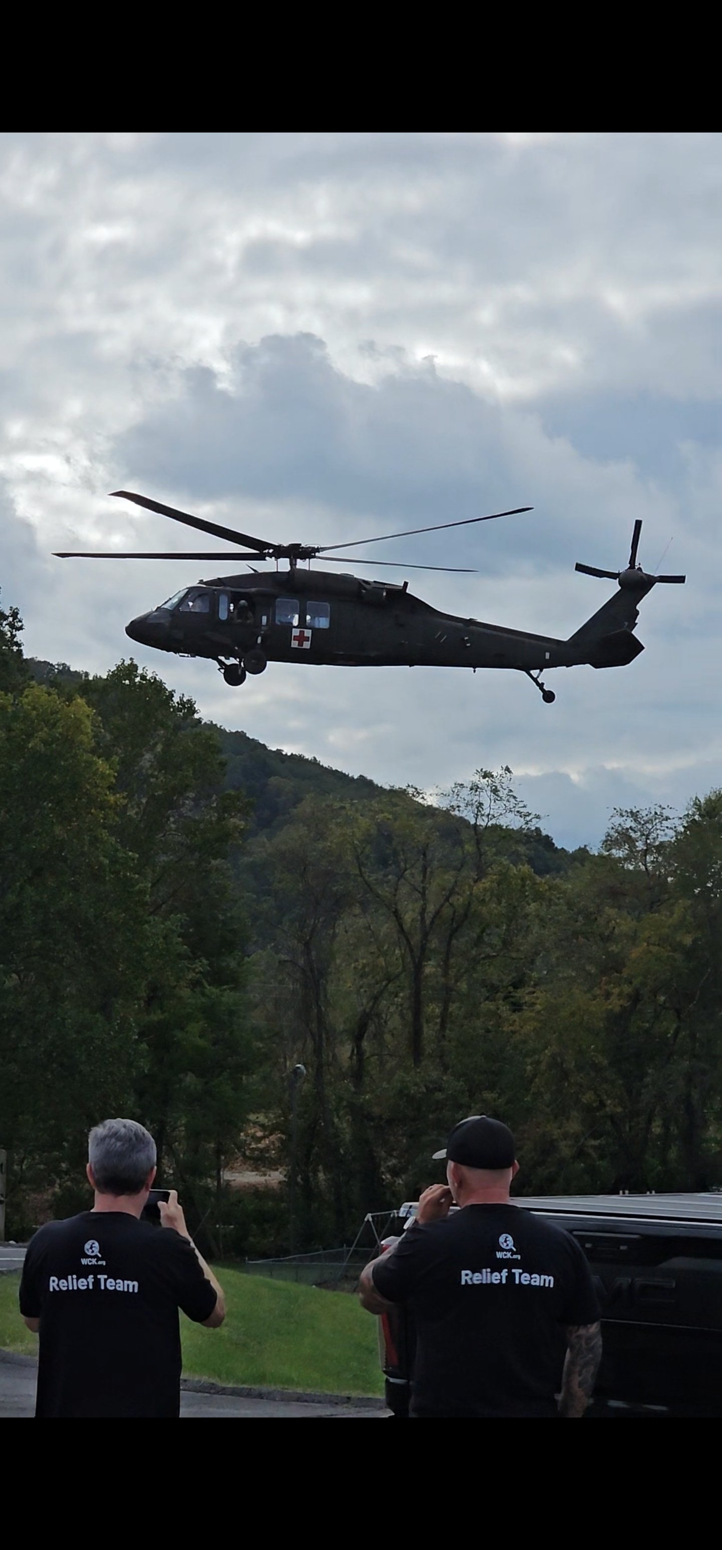 A helicopter flying in the sky above a wooded area, with two people in black shirts taking photos of it from the ground.