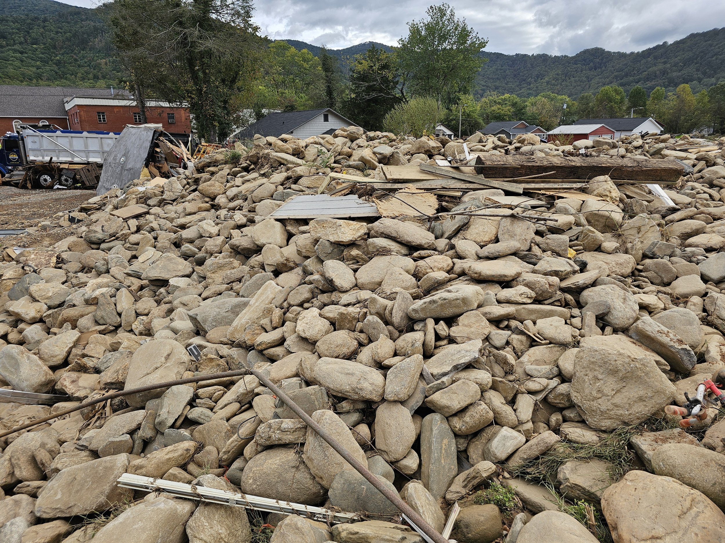 Large pile of rocks and debris from flood damage with damaged structures and scattered debris in a rural area.