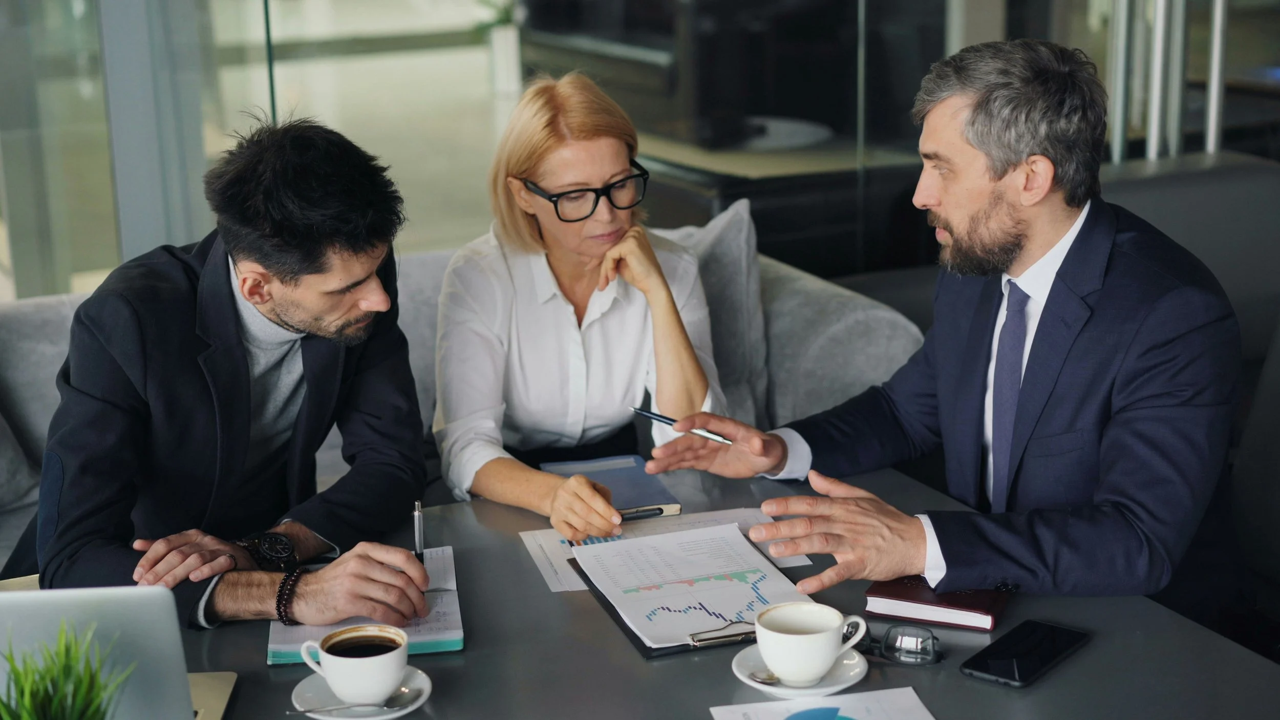 Three business professionals having a discussion at a table with documents, charts, and coffee cups.