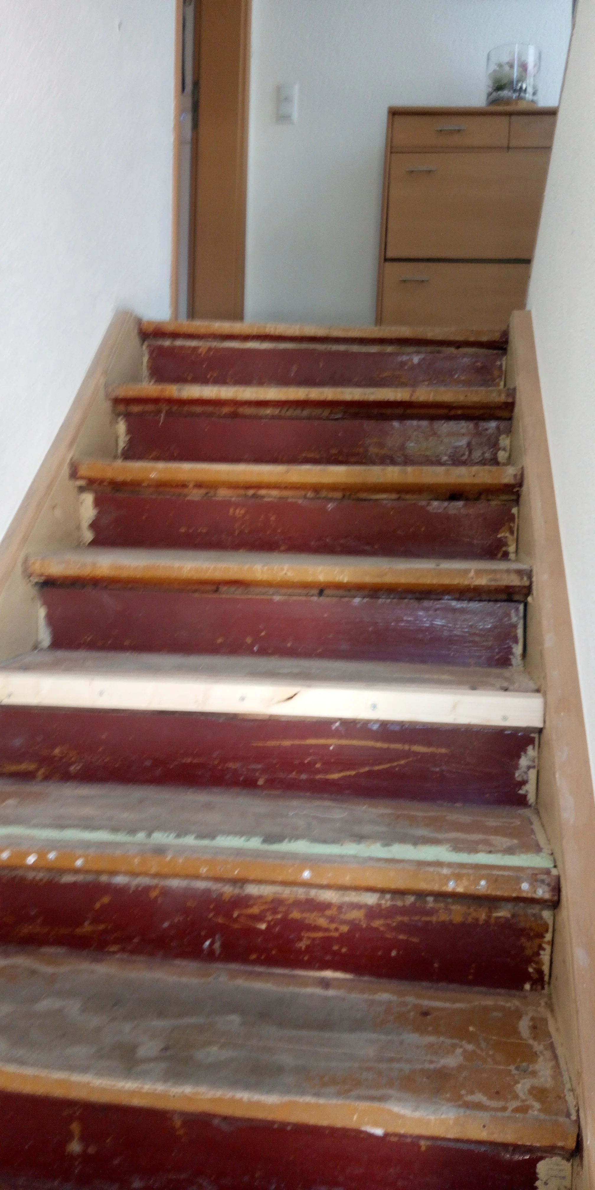 Innenraum eines Hauses mit abgenutzten Treppenstufen aus Holz, auf dem Flur steht eine Holzkommode mit einem Glastopf auf der Oberseite.