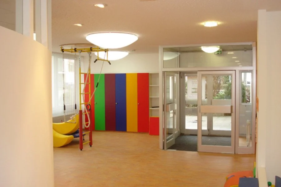 Innenraum eines Spielzimmers mit bunten Wandschränken, Klettergerüst, Rutschen und großen Fenstern.