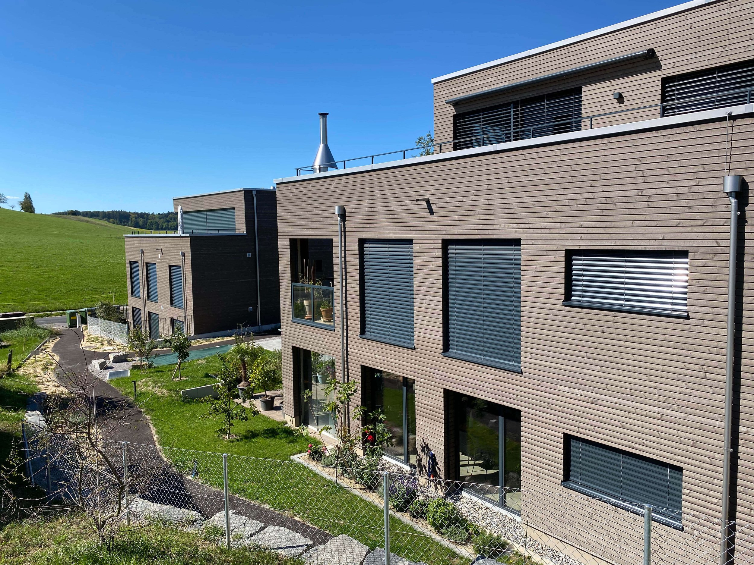 Moderne mehrstöckige Wohngebäude mit Holzfassaden, große Fenster mit Rollläden, Garten mit Wegen, Bäumen und Zierpflanzen, grüne Wiesen im Hintergrund, blauer Himmel.