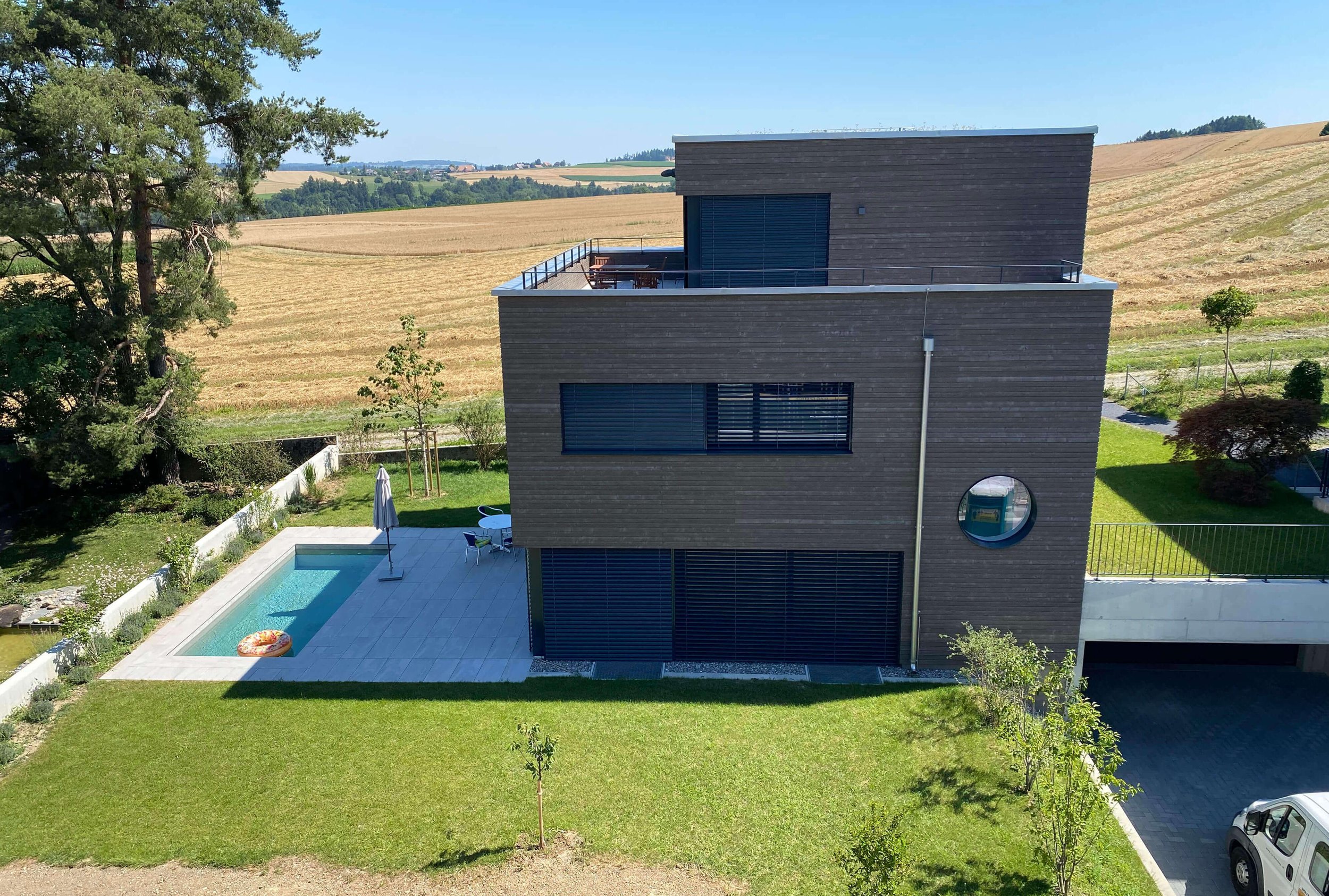 Moderne schwarze Villa mit Garten, Pool und Terrasse auf dem Land, umgeben von Feldern und Bäumen.