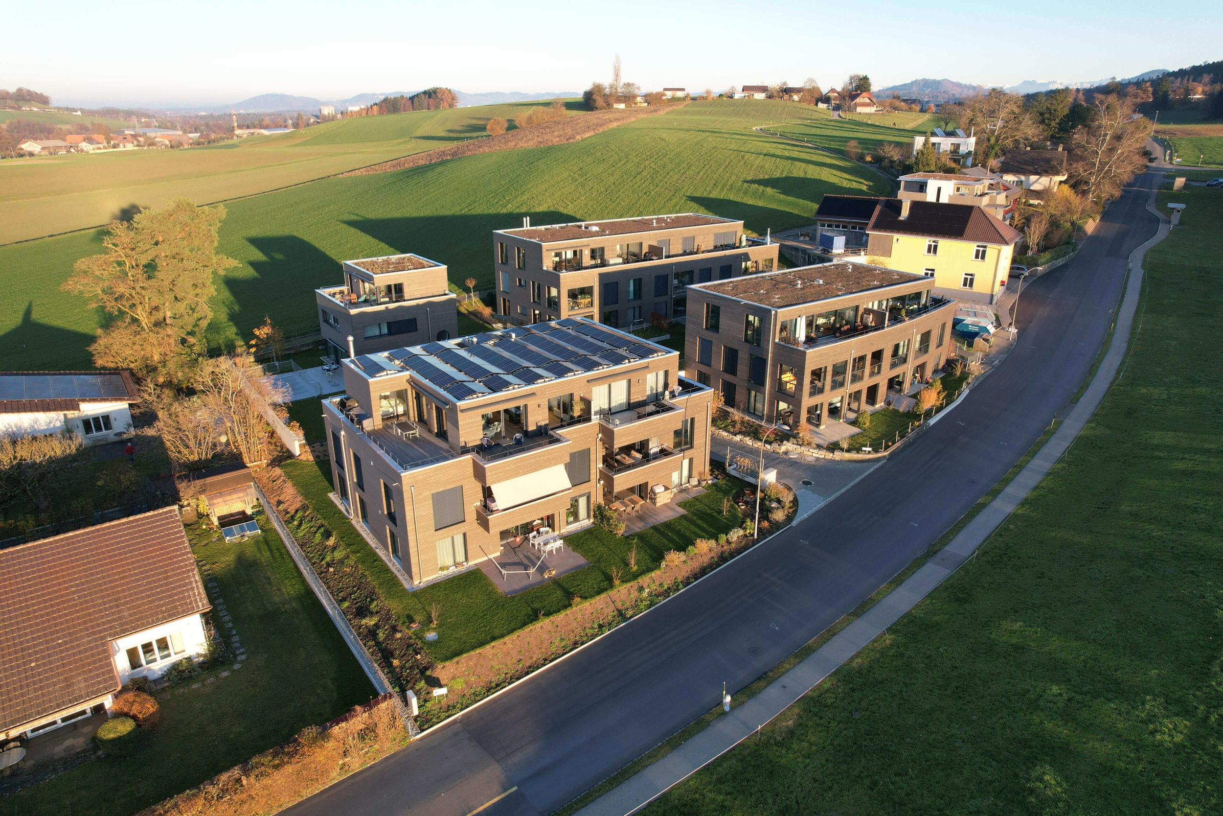 Mehrstöckige moderne Wohngebäude mit Solarpaneelen auf den Dächern, umgeben von Grünflächen und einer ruhigen Straße in einer ländlichen Gegend.