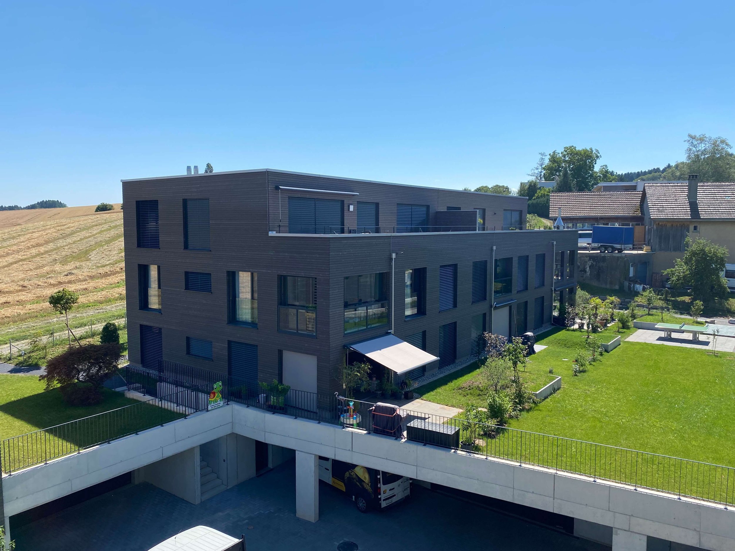 Mehrstöckiges modernes Wohngebäude mit großer Grünfläche, Bäumen und Gartenmöbeln.