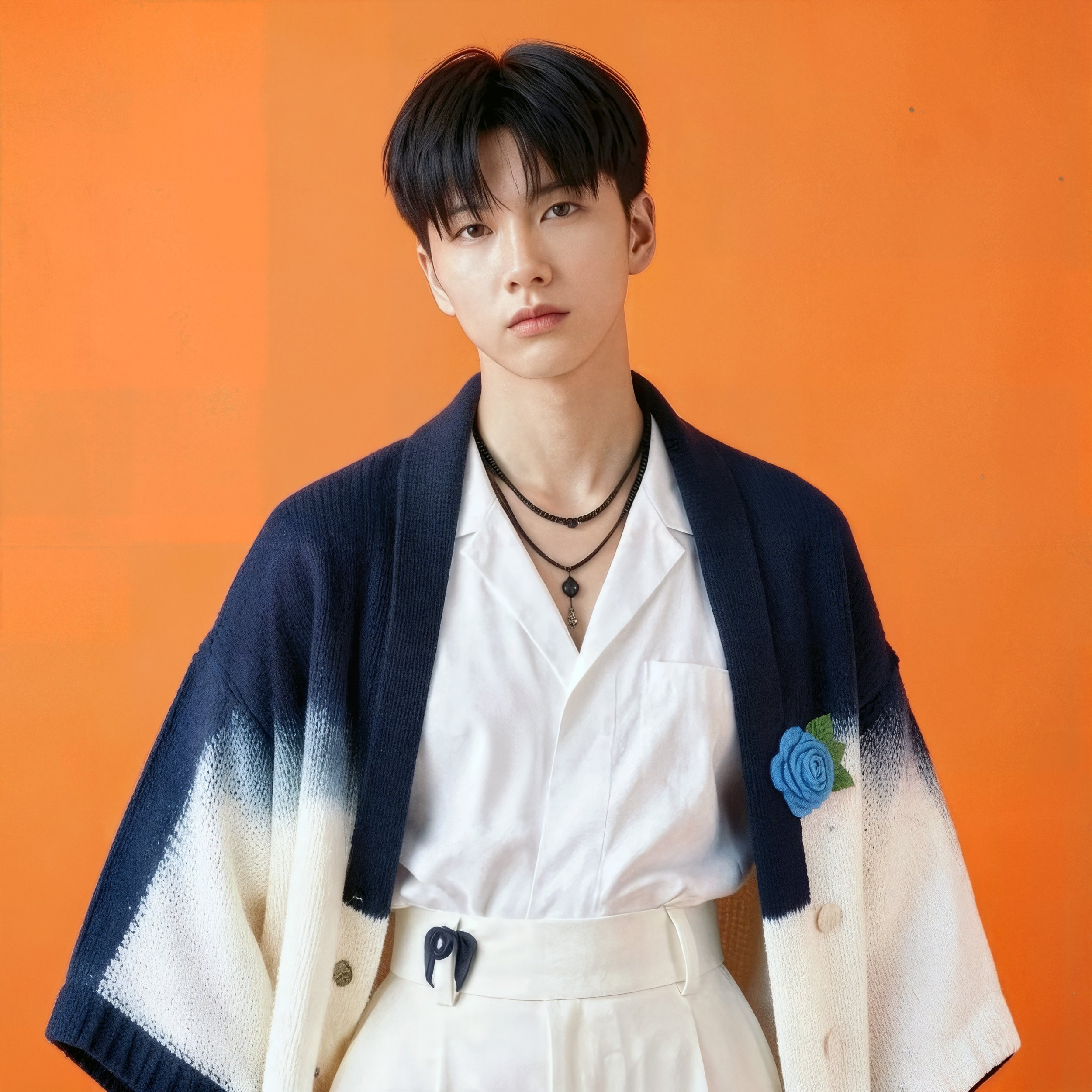 Young man with black hair wearing a white shirt, beige skirt, and a blue and white cardigan with a blue rose patch on the chest, against an orange background.