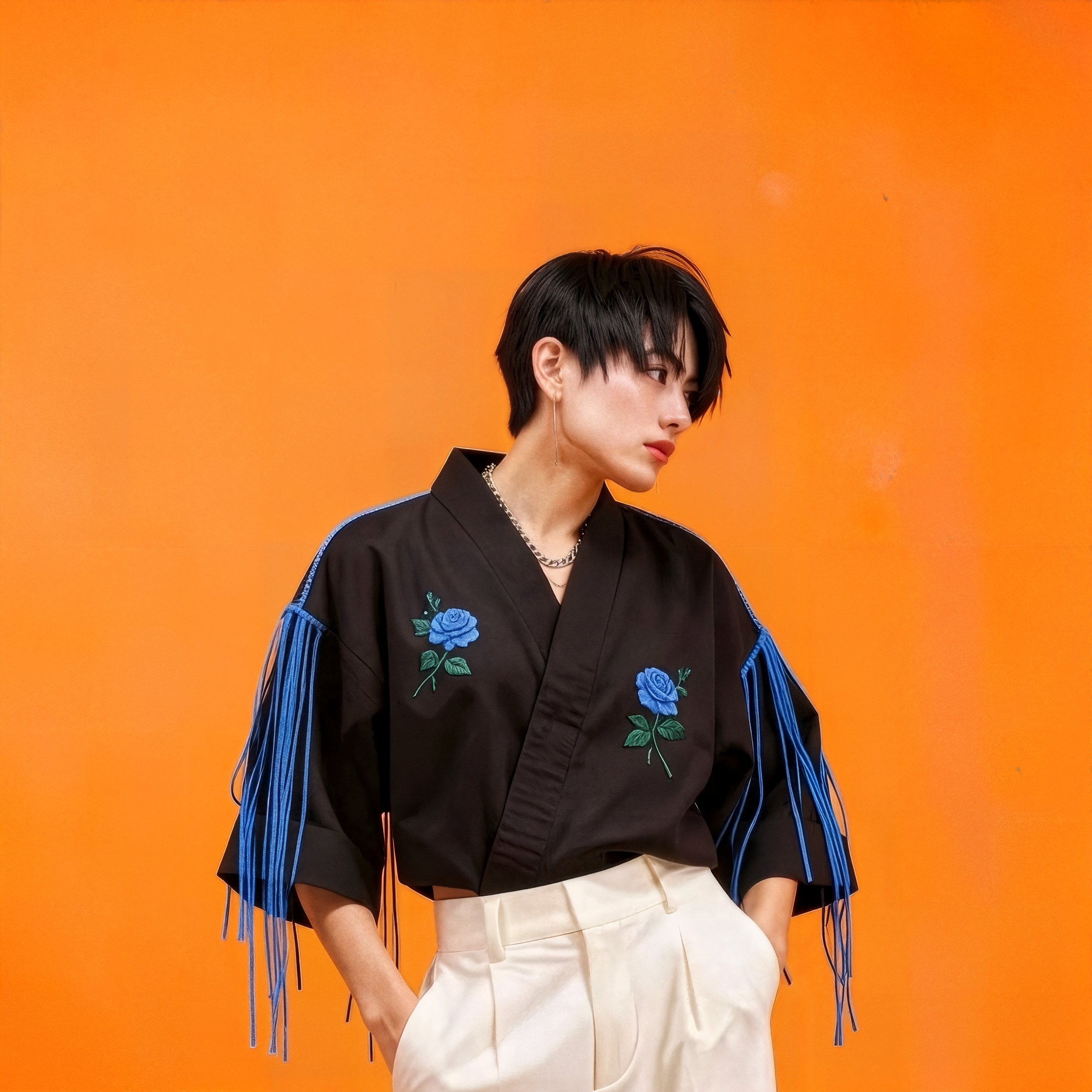 A woman wearing a black shirt with blue embroidered roses and blue fringe details on the sleeves, standing against an orange background.