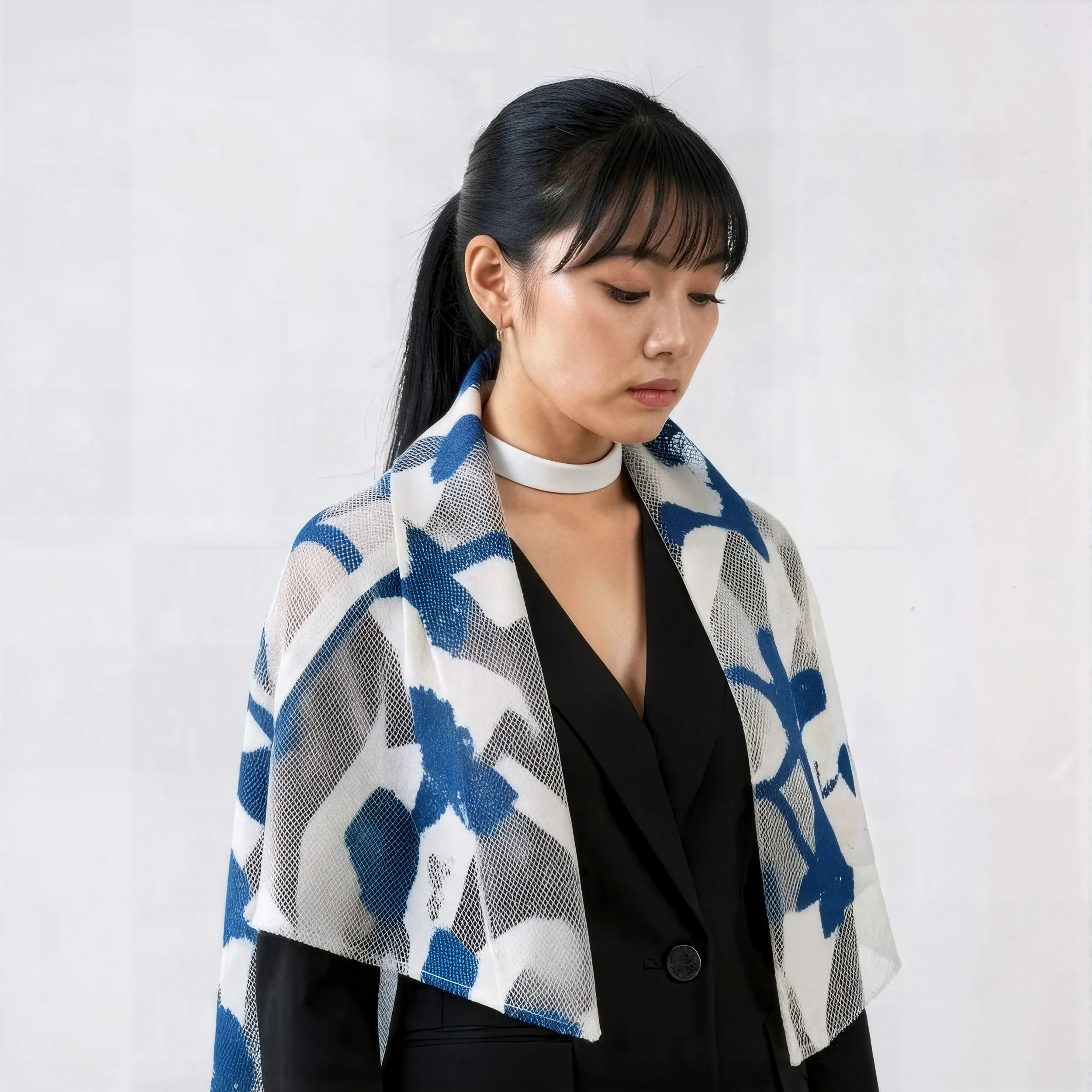 Young woman with black hair tied back, wearing a white choker, black blazer, and a sheer shawl with blue and white leaf pattern, standing against a plain white background.