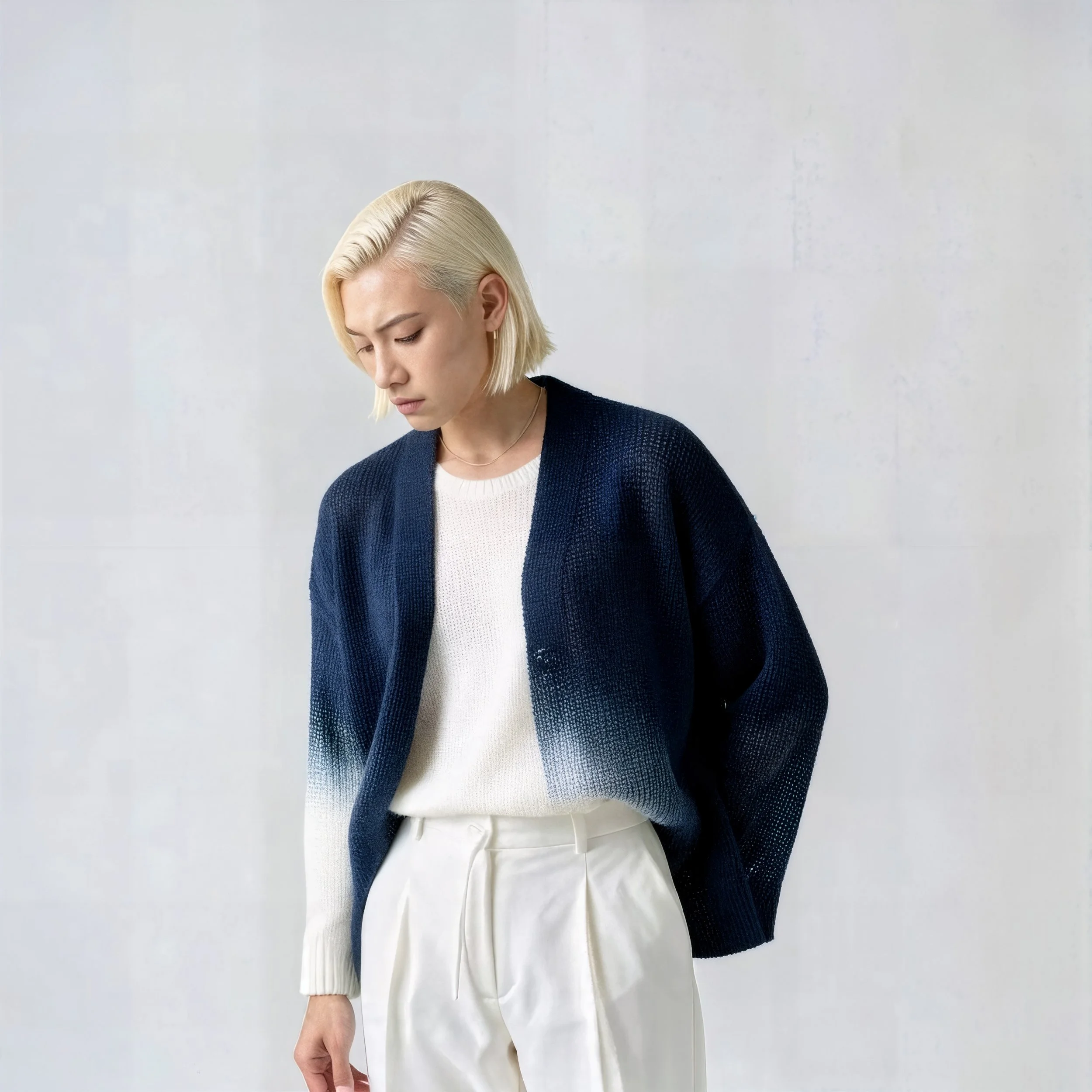 A woman with short blond hair looking down, wearing a navy blue ombre cardigan, a white sweater, and white pants, standing against a light gray wall.