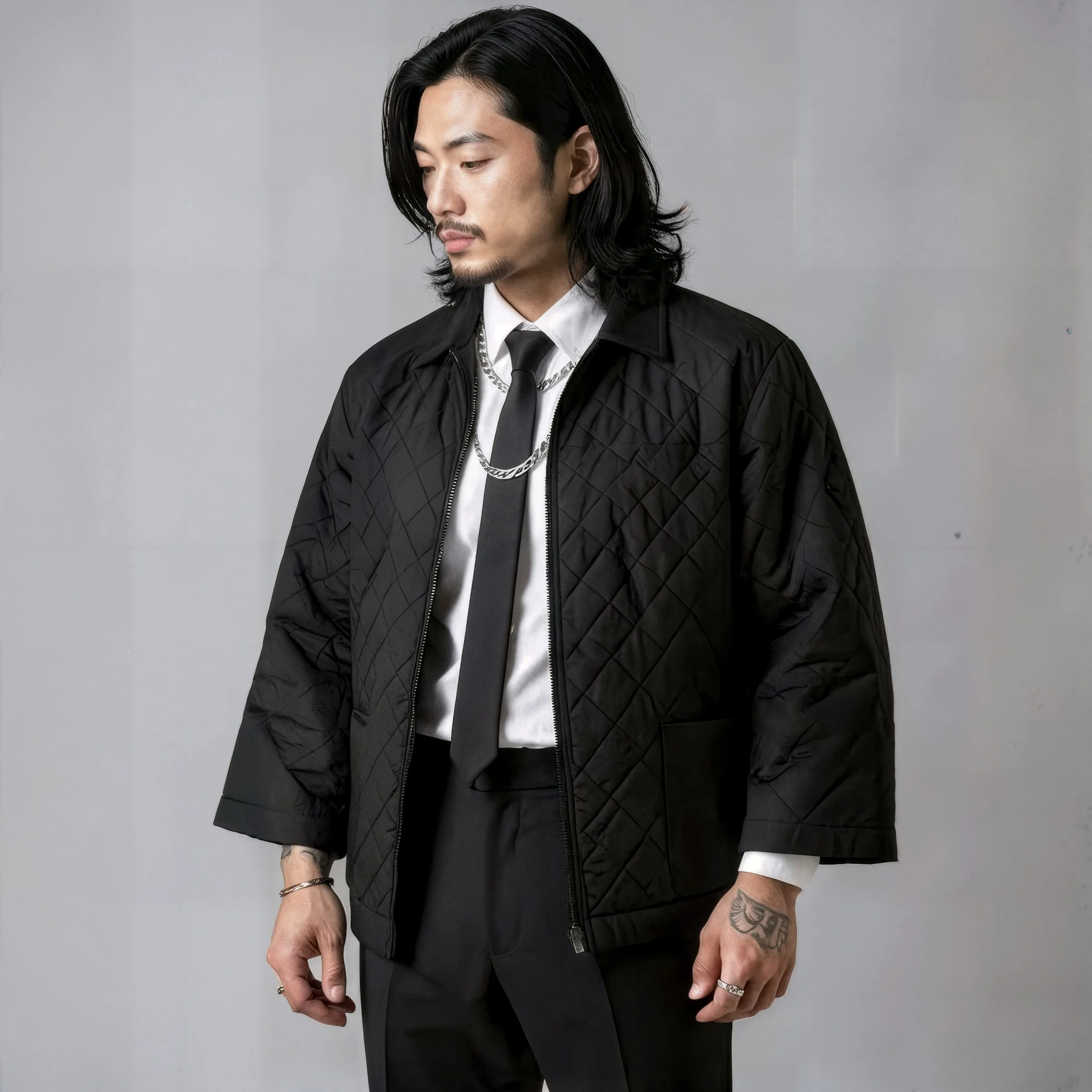A man with long black hair dressed in a black quilted jacket, white shirt, black tie, and accessories, standing against a plain background.