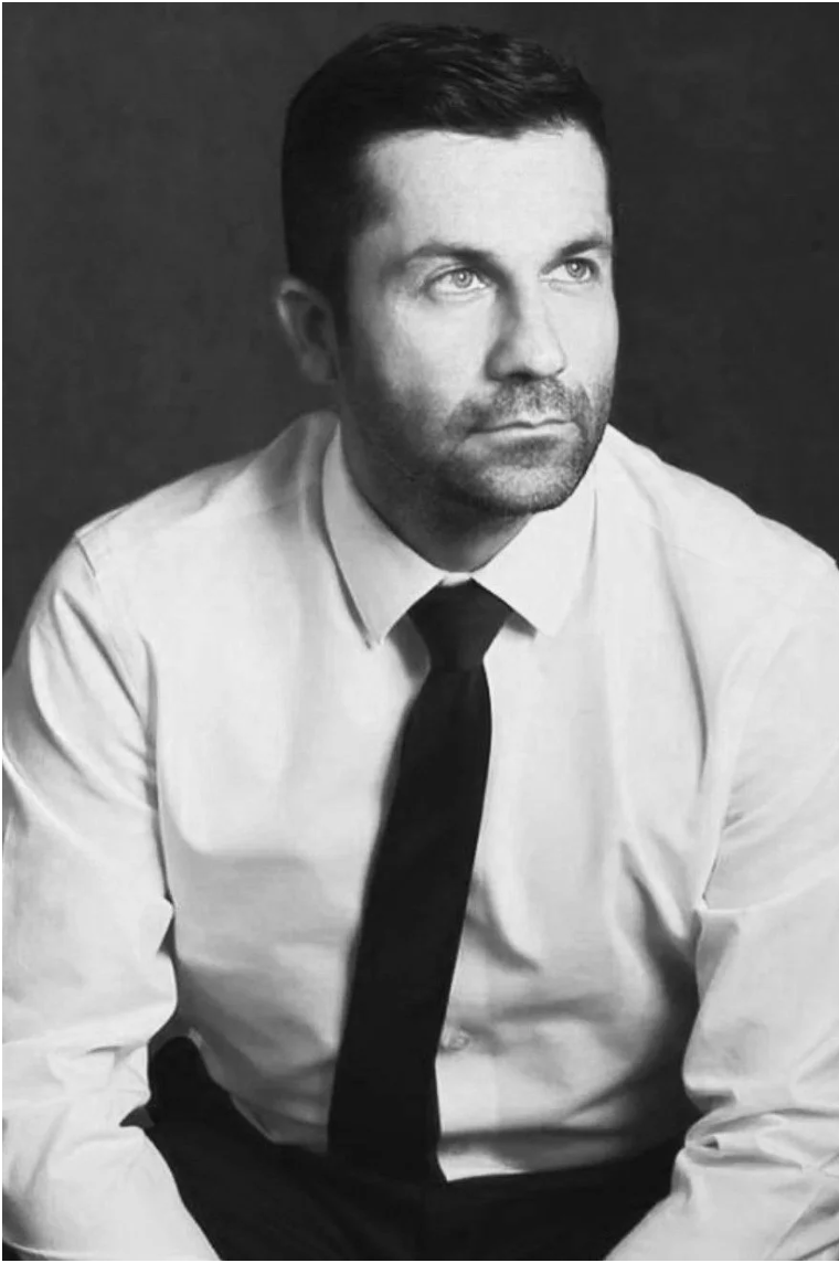 Black and white portrait of a man with short dark hair, wearing a white shirt and dark tie, looking thoughtfully to the side against a dark background.