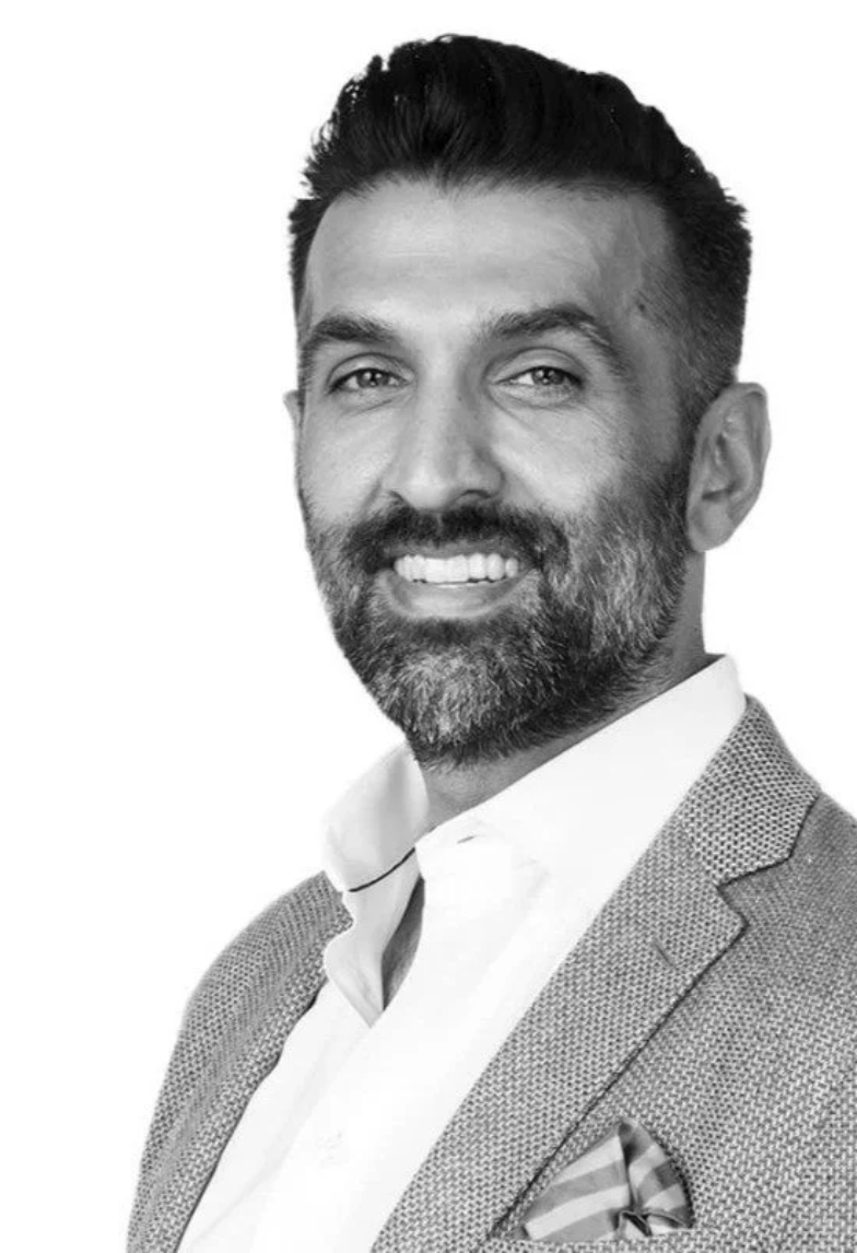 A black-and-white photo of a smiling man with dark hair, a beard, and mustache, wearing a suit jacket, dress shirt, and pocket square.